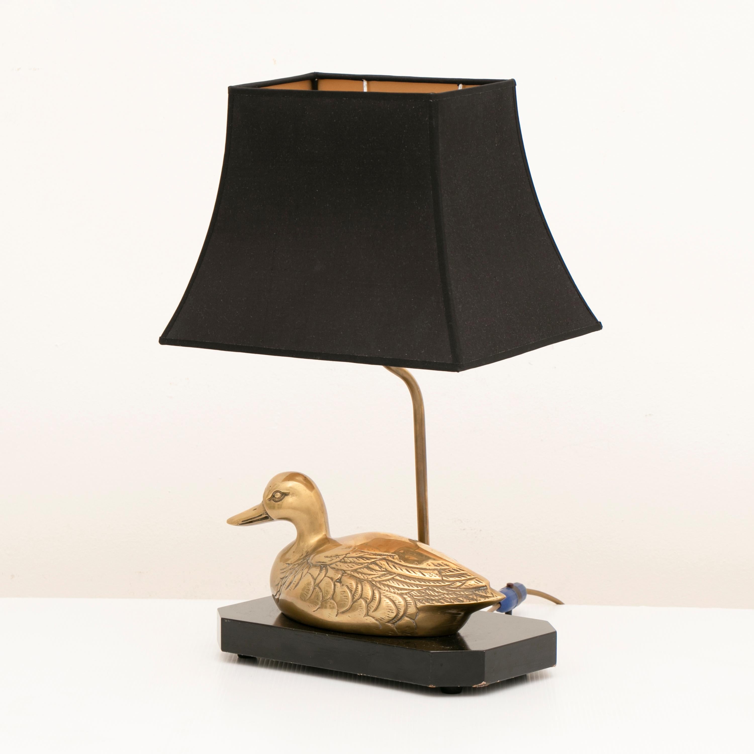 1970s vintage Hollywood Regency brass duck table lamp which is mounted on a black lacquered wooden base in the style of Maison Jensen. The brand new shade is held in place by a brass rod which is fixed to the base at the back of the duck. A single