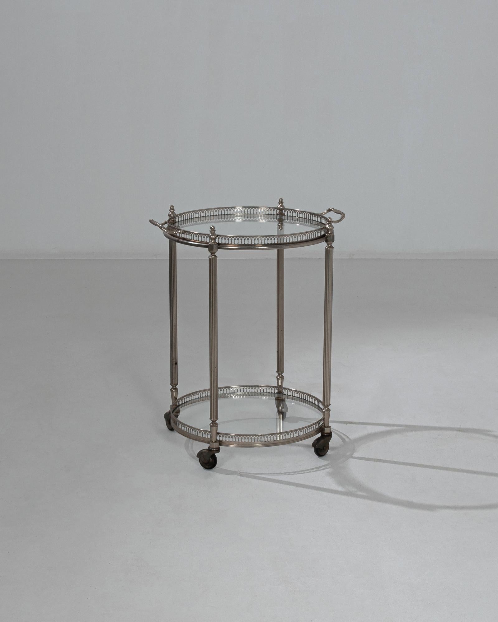 This metal bar cart was made in France, circa 1970, and imparts a genteel elegance with its refined ornamentation and polished patina. Fluted legs topped with spires support both tiers, crowned with a circle of dainty arched spines. Contoured