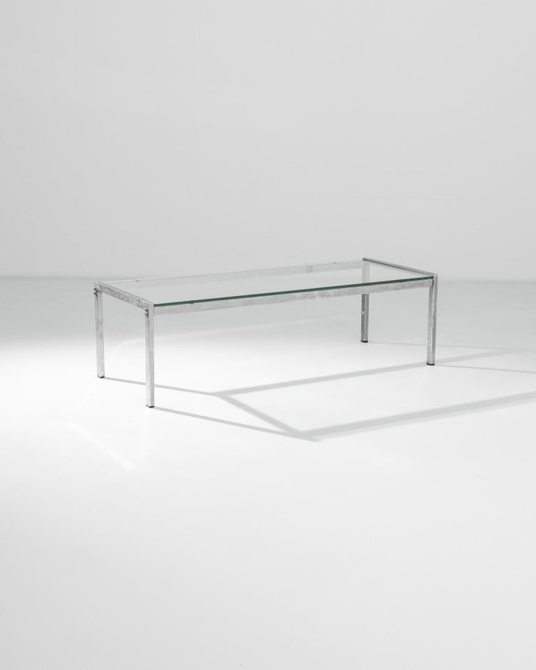 A metal coffee table with glass top from France, produced circa 1970. A minimalist, low-slung coffee table featuring a slender metallic frame and thick, glass table top. The clean lines of the frame are an exemplar of the future oriented design of
