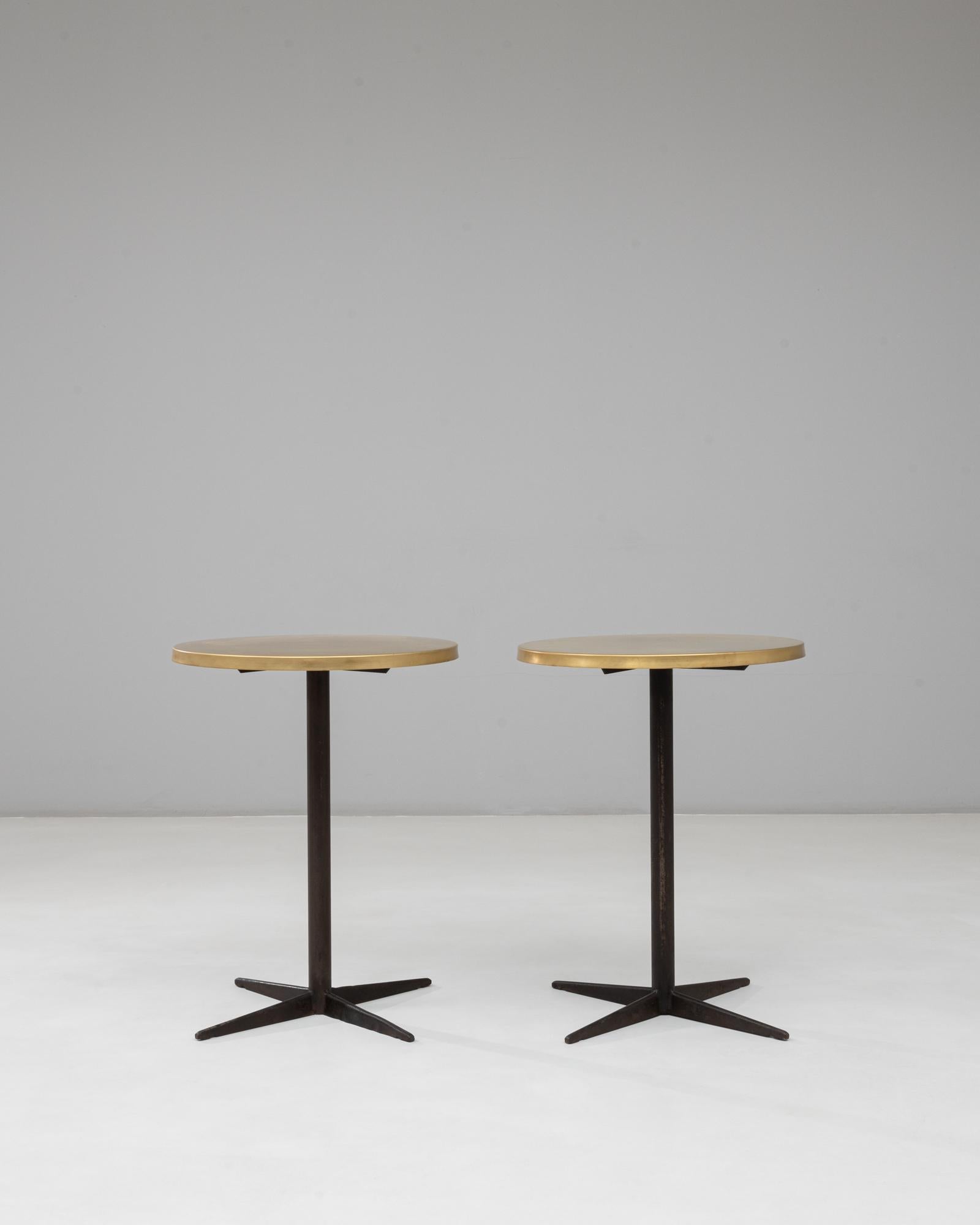 This 1970s French pair of bistro tables embodies the sleek sophistication of mid-century design. Each table features a solid iron base with a four-point pedestal form, offering stability and industrial appeal. The round brass tops add a luxurious