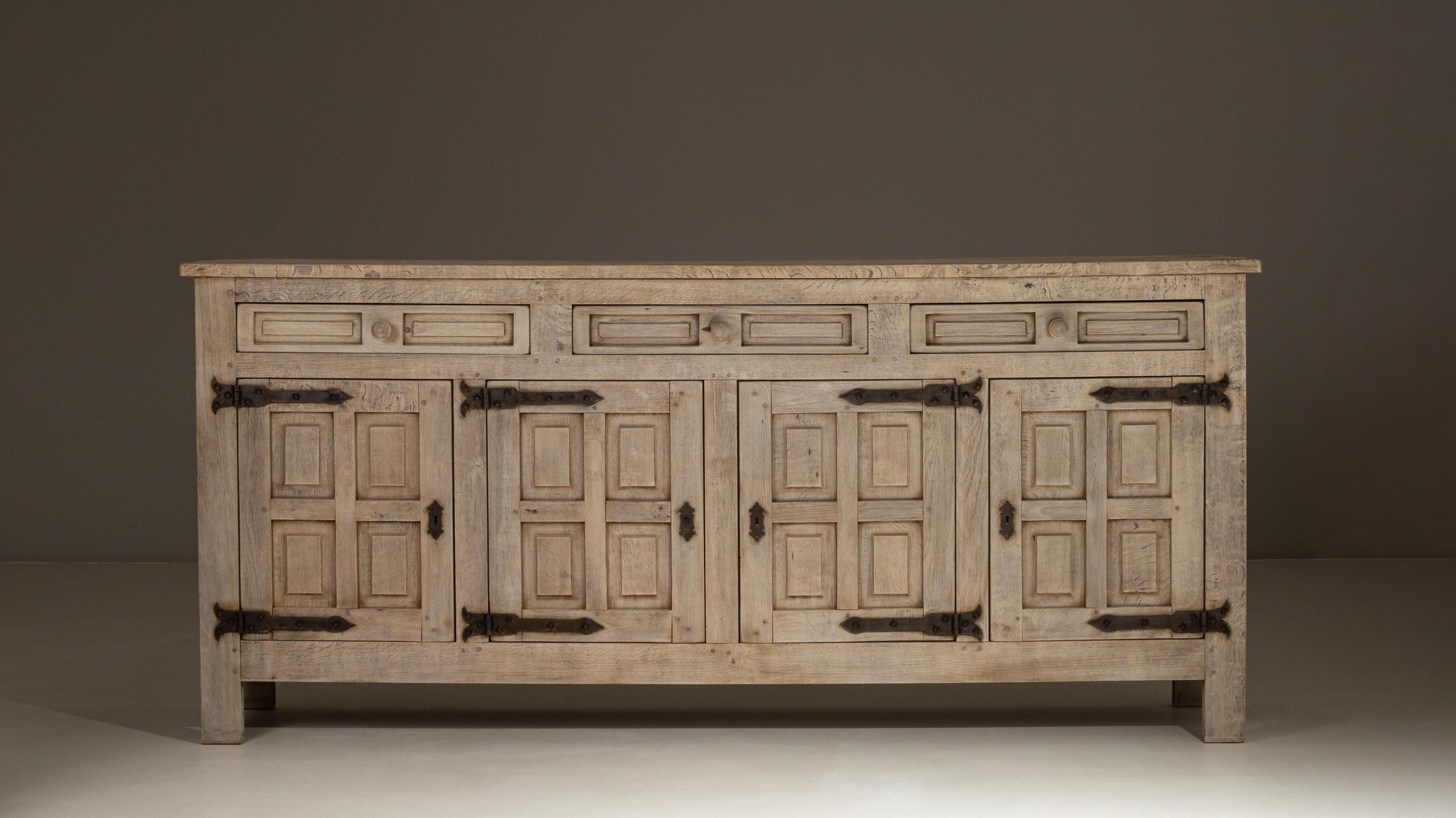 This 1970s French Primitive oak buffet features a substantial, architectural presence defined by its paneled doors, robust frame, and beautifully weathered natural finish. Crafted from solid oak, the piece highlights the rustic character of the wood