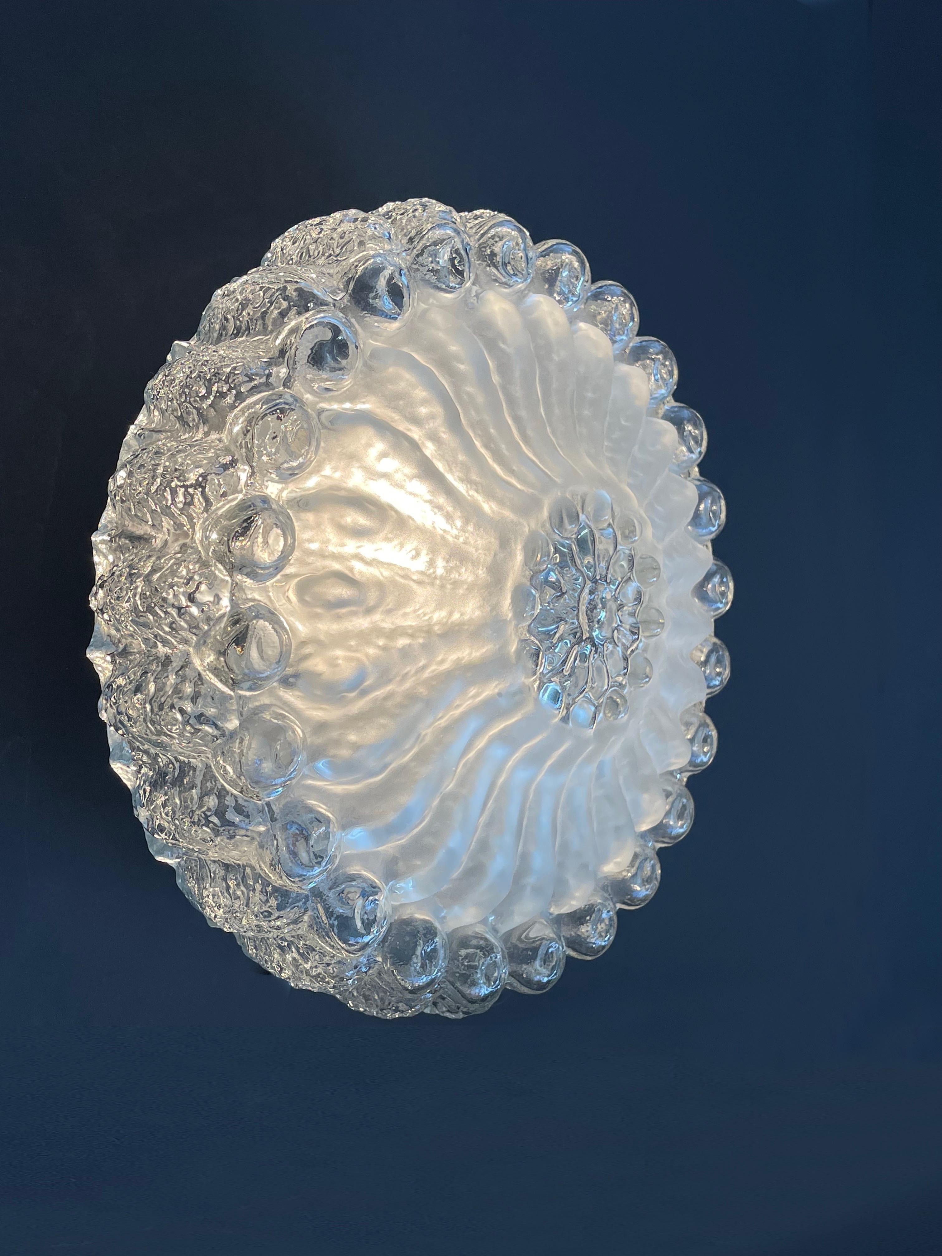 Beeindruckende opulente Decken- oder Wandleuchte mit bündigem Sockel, hergestellt in Deutschland um 1965-1975.
Außen klobiges, leicht steiniges, klares Blasenglas, in der Mitte bildet Milchglas die Blütenblätter.
Durch diesen Kontrast entsteht ein