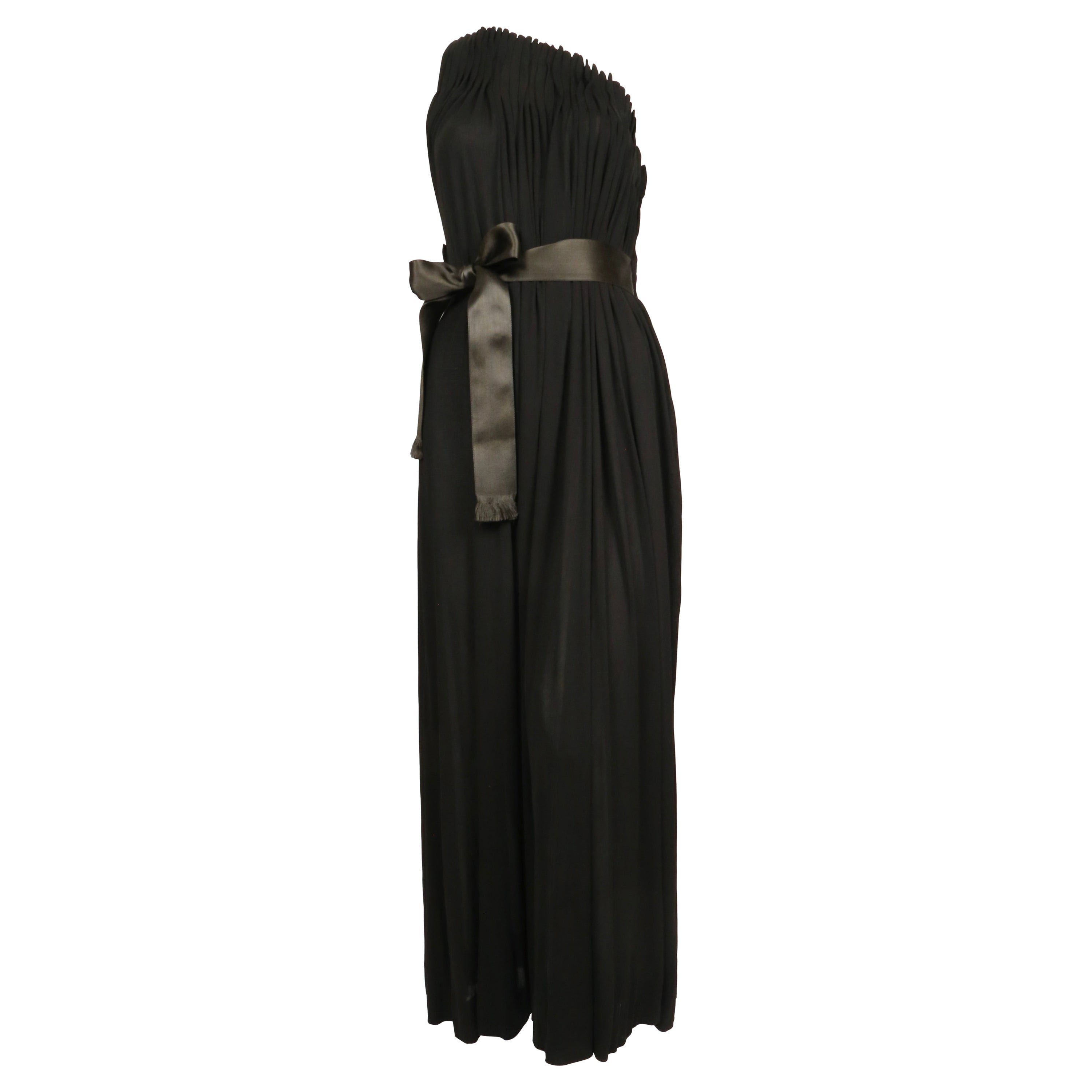 Impresionante vestido negro azabache de jersey de seda con corpiño plisado acanalado de James Galanos de los años setenta. El vestido tiene una silueta muy marcada con un singular escote alto asimétrico con un único tirante fino en la espalda.  A