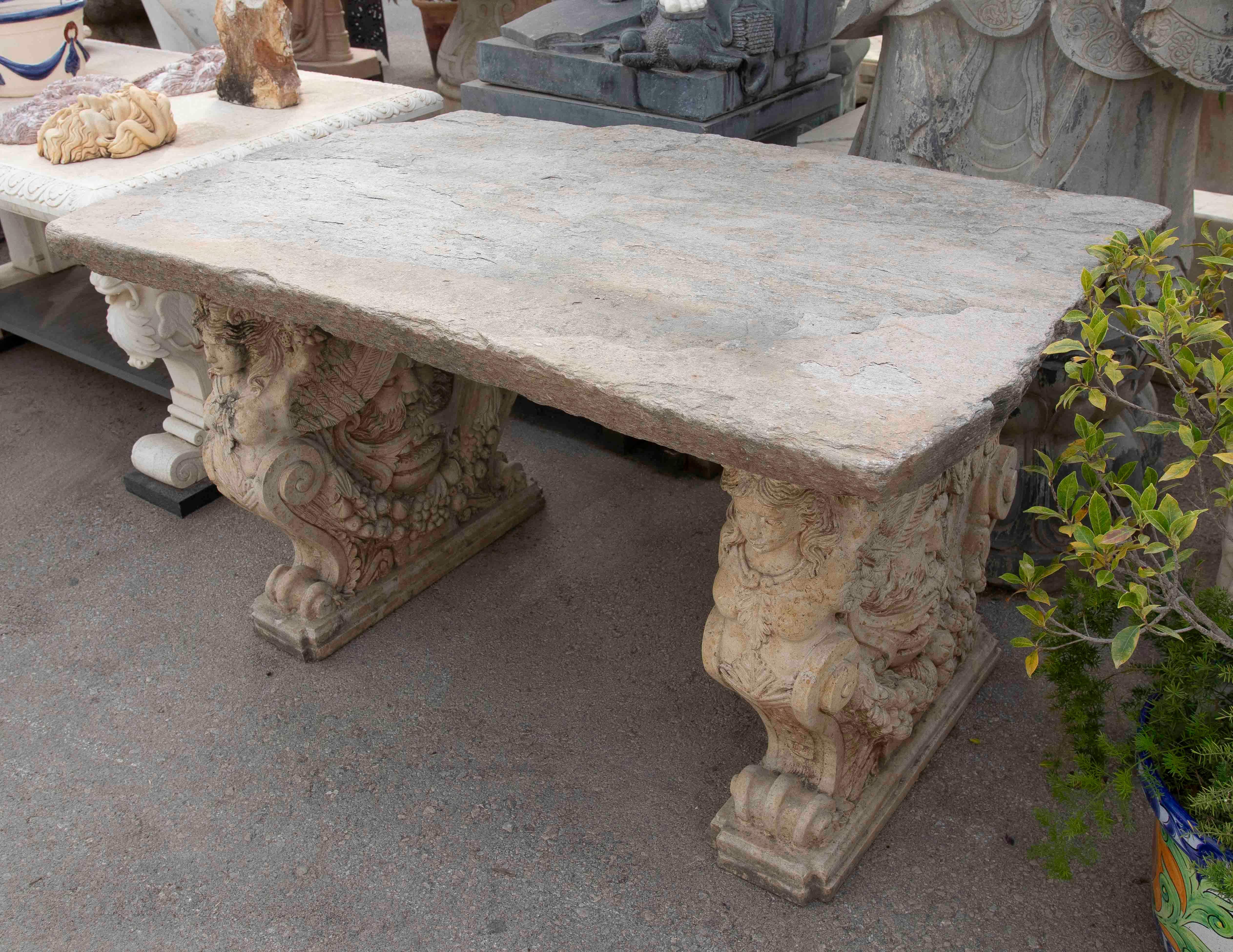 An impressive garden table made of hand-carved stone, with a striking and sculptural character. The organically shaped tabletop with a rustic finish rests on two spectacular supports decorated with mythological figures and plant motifs, crafted with