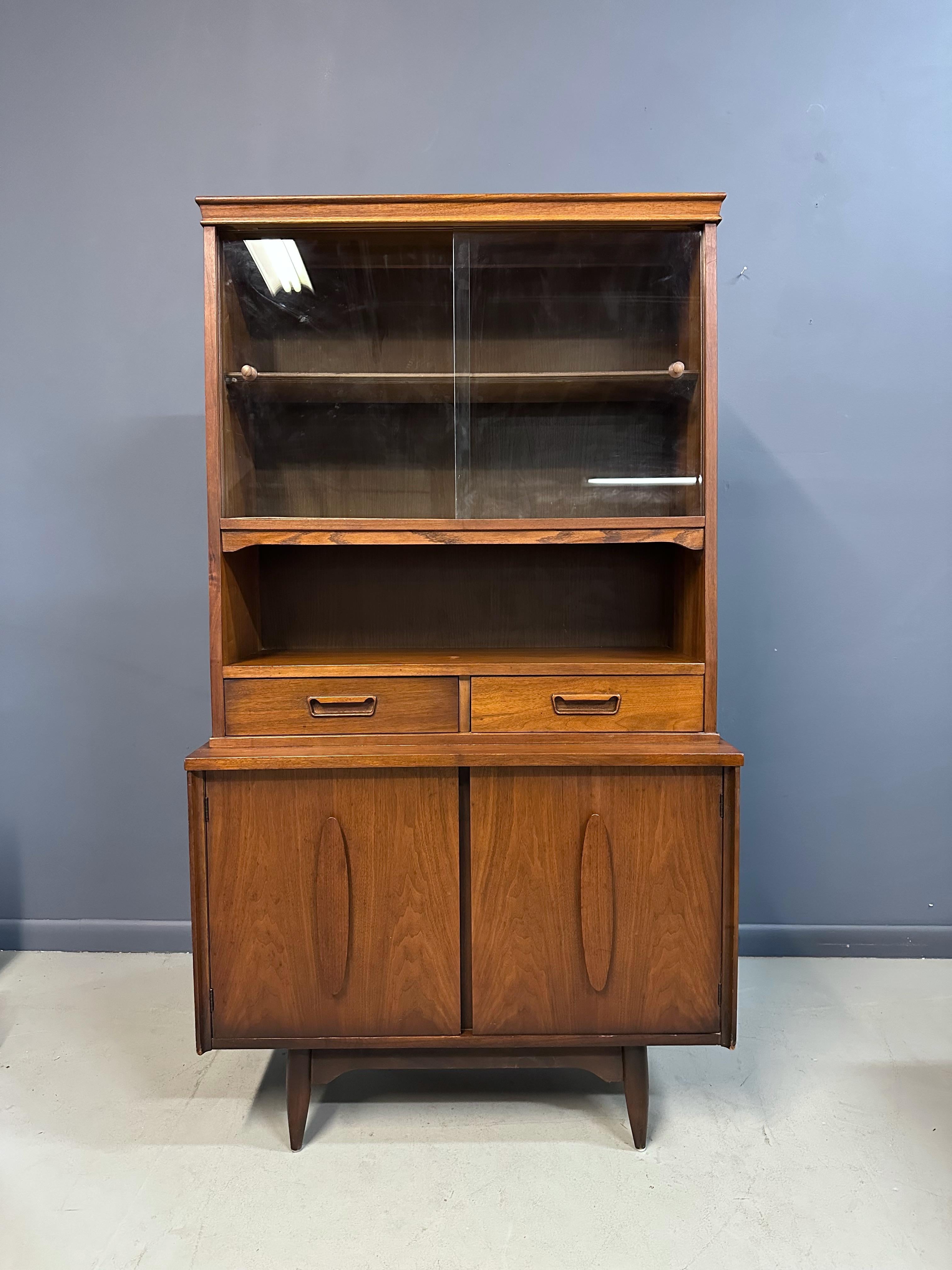 This 1970s Garrison Mid Century walnut china cabinet exemplifies timeless craftsmanship and elegant design. Featuring two spacious doors, it offers ample storage for your fine china or collectibles while showcasing the rich walnut finish