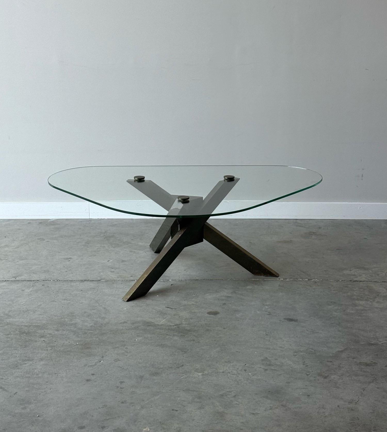 A unique 1970s brass and glass coffee table. A sculptural brutalist base. Triangular center with three long posts, wonderful patina. Sitting atop the base is a large triangular shaped glass top. Screws with rounded brass caps lock the glass top into