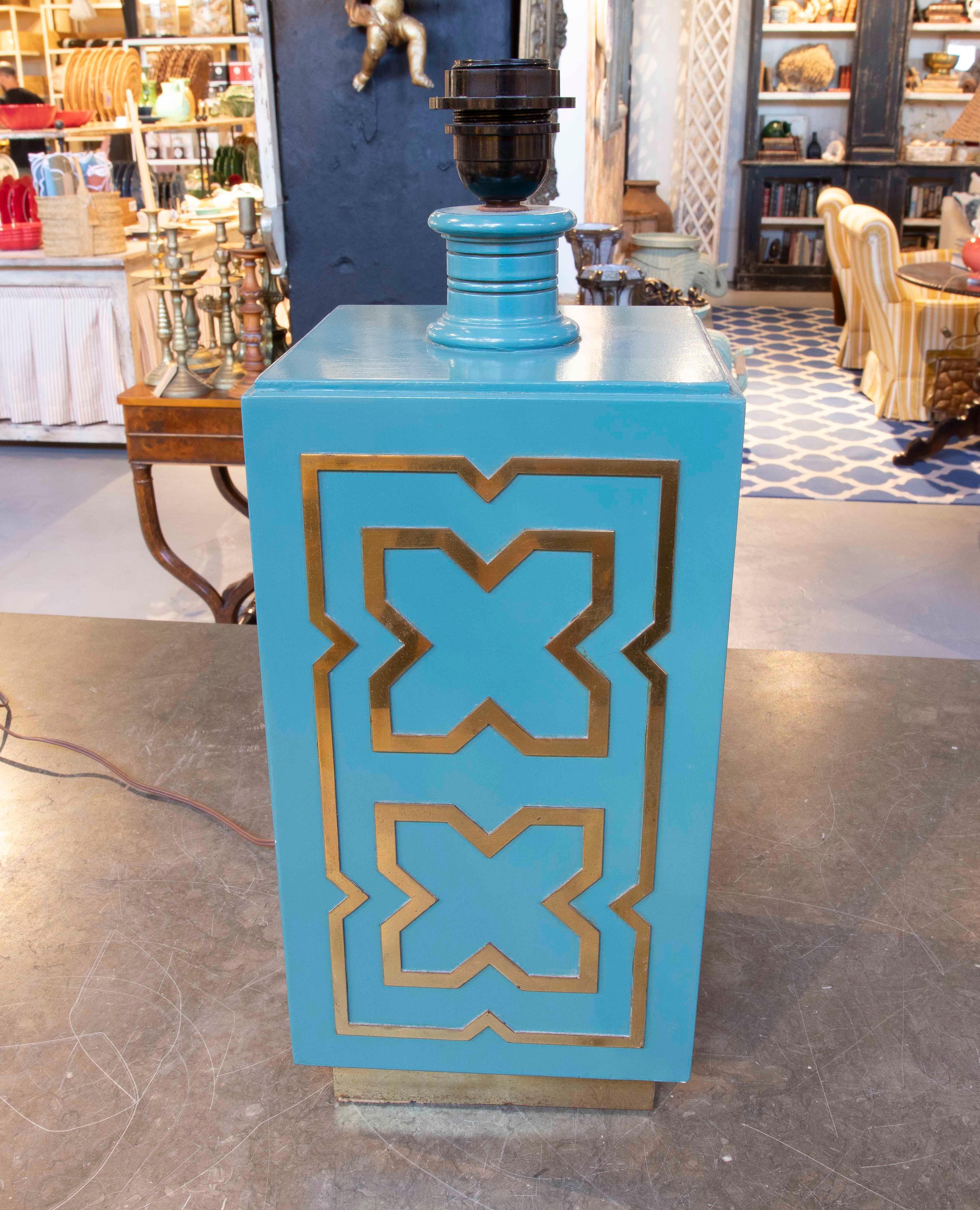 Lampe de table géométrique, fabriquée en bois laqué turquoise avec des détails décoratifs en laiton doré gaufré, inspirés des motifs mauresques ou mudéjars. Sa base rectangulaire élancée et sa couleur vibrante en font une pièce d'accentuation idéale