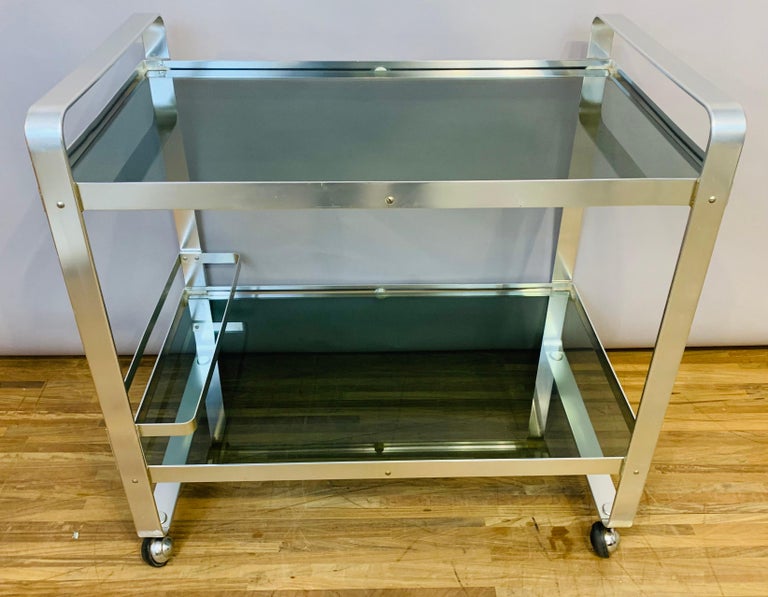1970s Space-age chrome and smoked glass drinks trolley or bar cart manufactured by Av Handwerk in Germany. The trolley is constructed with an aluminium metal frame and smoked dark green glass on castor wheels. A bottle holder which can slide up and