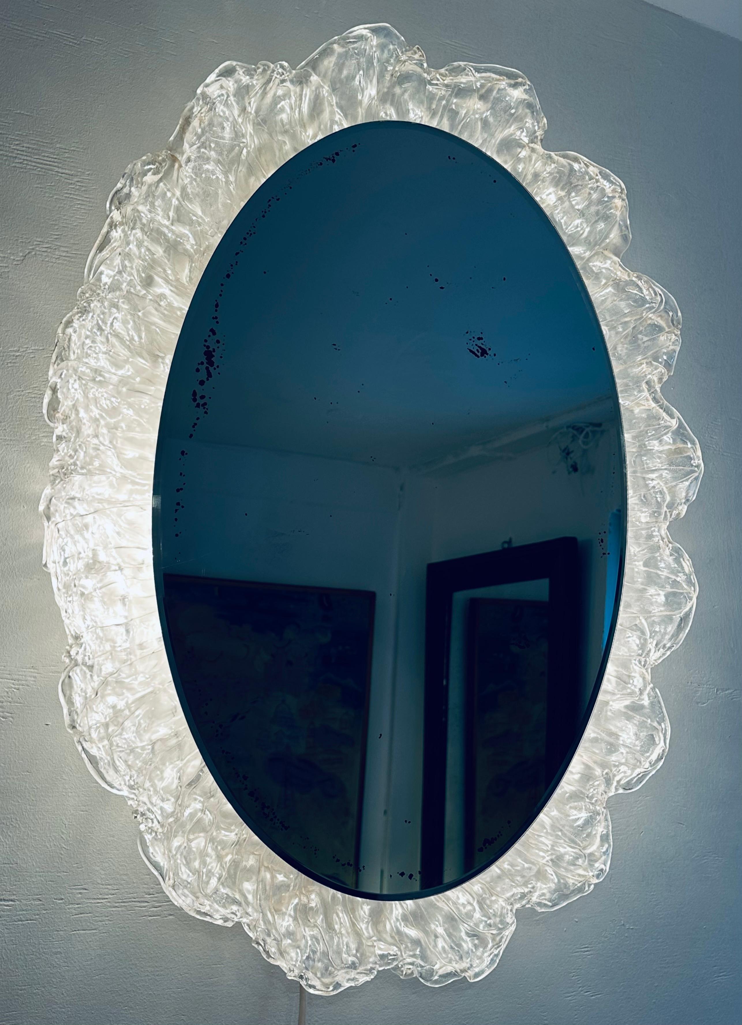 1970s German Hillebrand Leuchten illuminated oval back lit wall mirror.  The mirror sits within an acrylic flower-shaped frame with inward turning petals surrounding it.  It also resembles flames around an oval sun.  The frame holds three lightbulbs