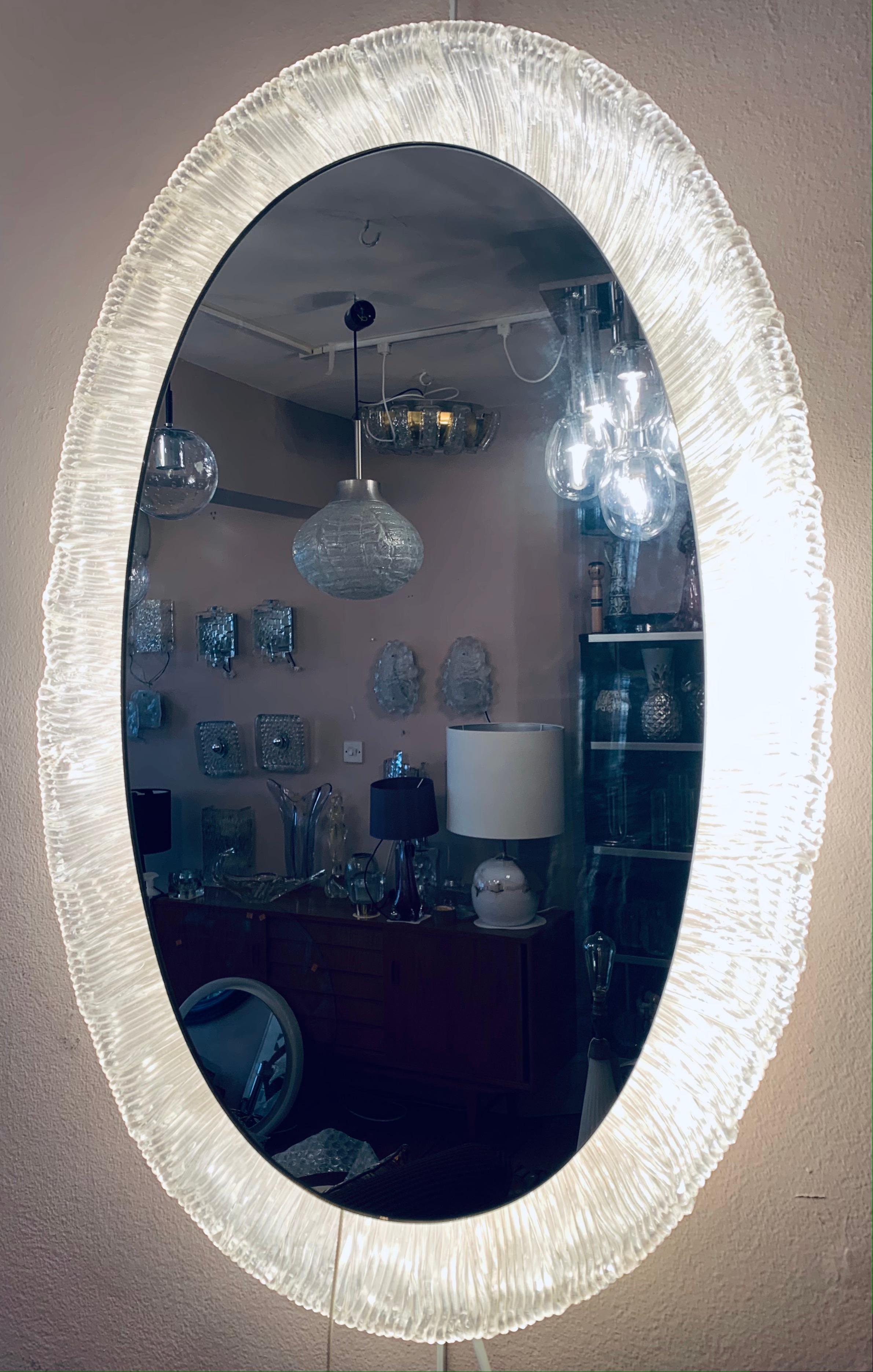 1970s German, Hillebrand lighting, illuminated, backlit, lucite-framed, oval, mirrored-glass wall mirror. The glass mirror is surrounded by a frosted-iced lucite frame which is screwed onto it. The frame holds 4 small screw in E14 light bulbs. I