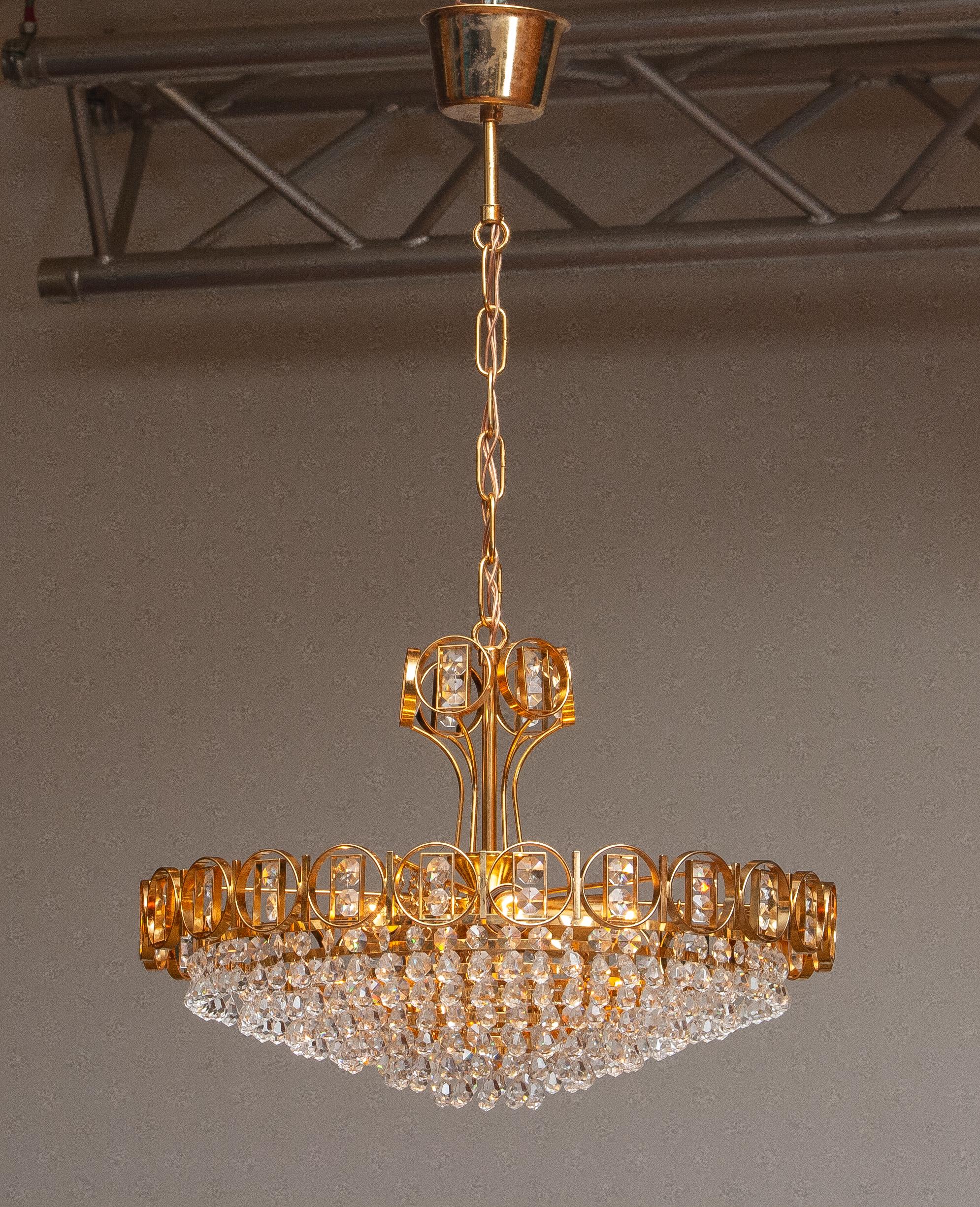 Magnifique lustre en laiton doré serti de cristaux facettés réalisé par Palwa, Allemagne, 1970.
Ce lustre est composé d'une couronne plaquée or avec à l'intérieur six anneaux tressés en or et sertis de cristaux et, au sommet, d'une couronne tressée