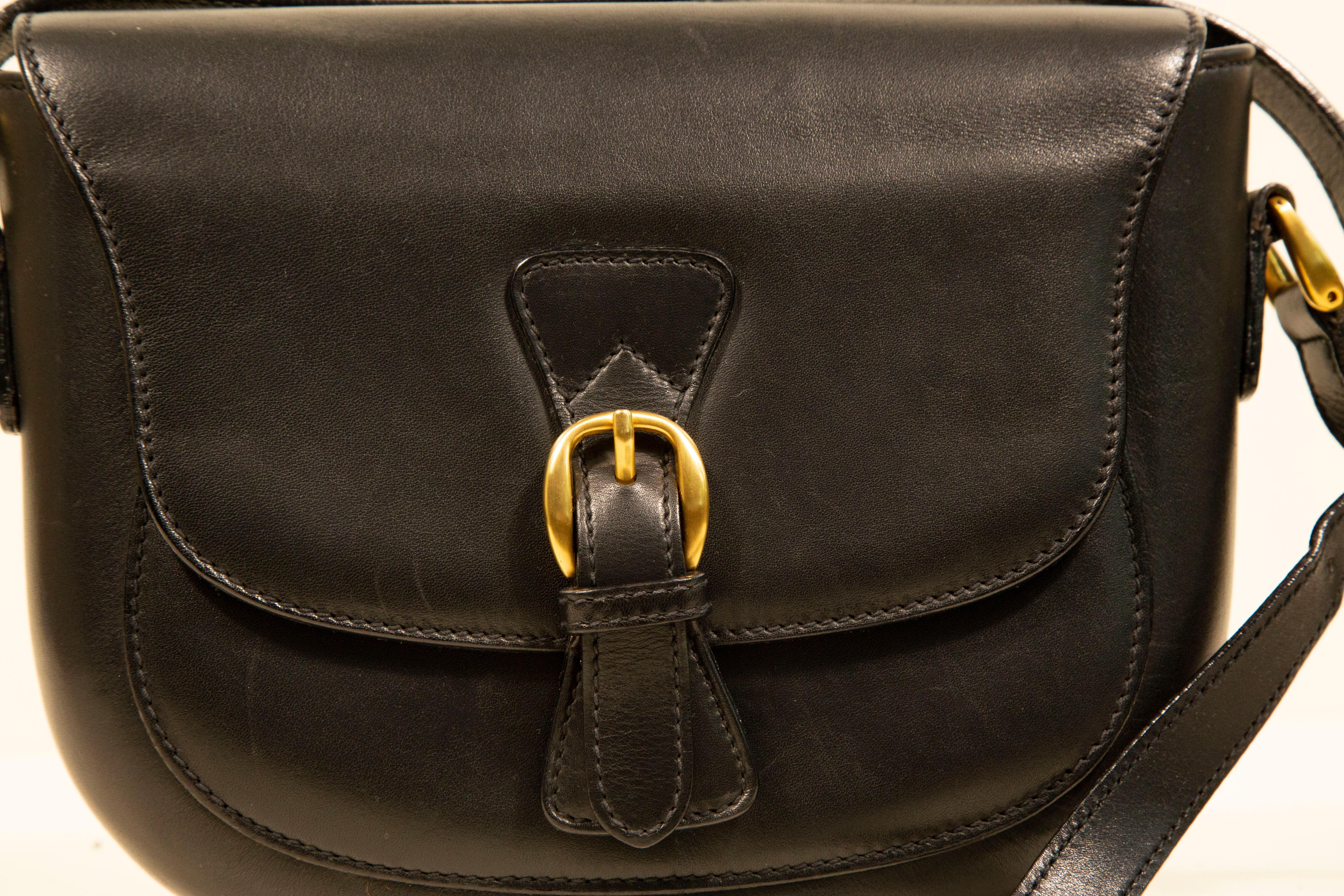 A vintage Gucci saddle bag/ crossbody bag made of black leather with gold toned hardware. The interior is lined with black suede, and next to the major compartment it features one side pocket with a zipper. The shoulder strap is in length