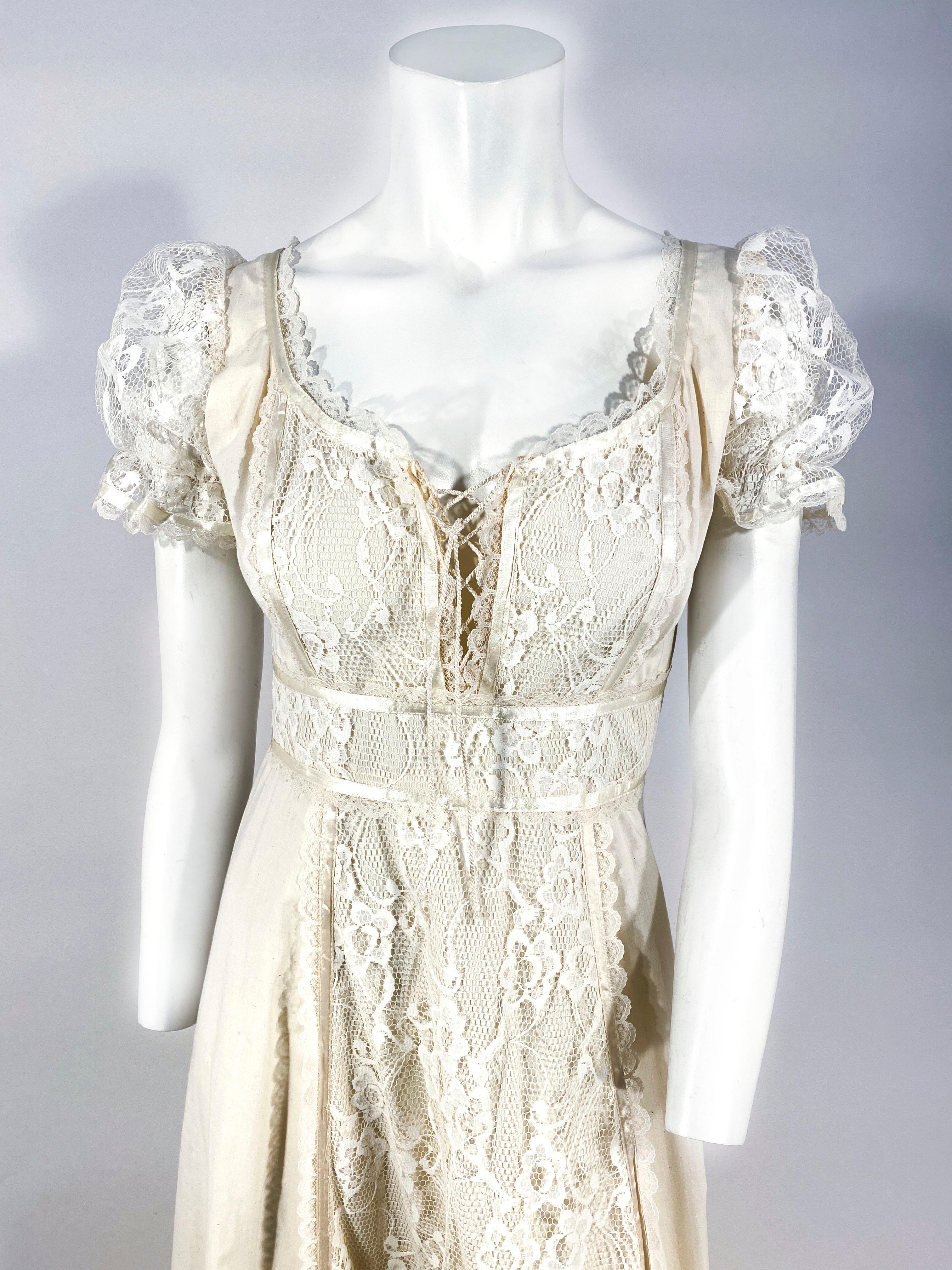 1970s Gunne Sax cotton cottage dress adorned with machine lace panels along the full-length skirt, a lace up bodice, sheer lace puff sleeves, satin ribbon trim, and an applied sash that ties in the back of the dress. There is also wide ruffled hem,