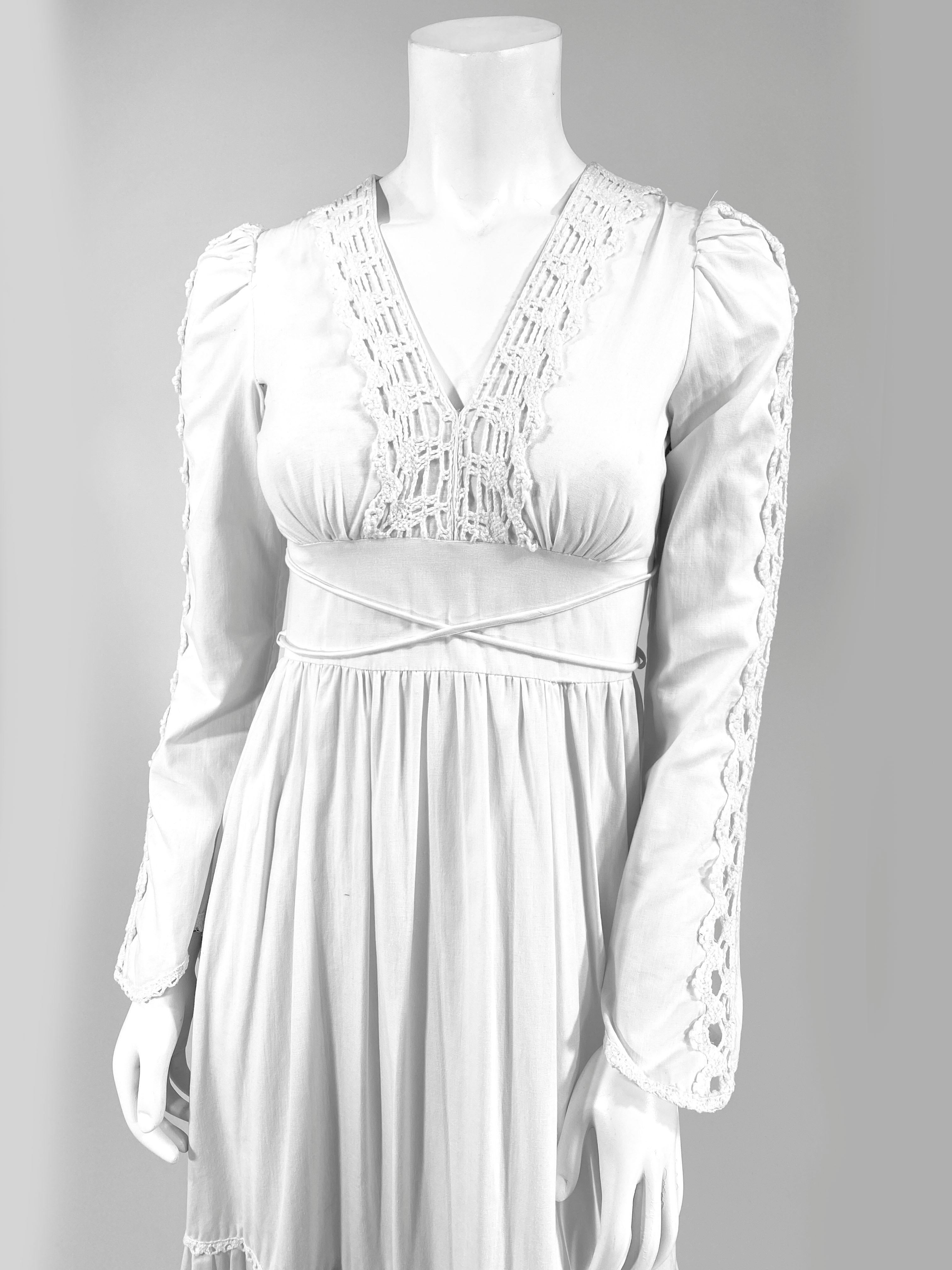 1970s soft cream colored cotton Gunne Sax Full length granny style dress with machine crochet trim along the full sleeves, deep v-neckline, and wide skirt ruffles. At the was it there is a criss-cross tie.