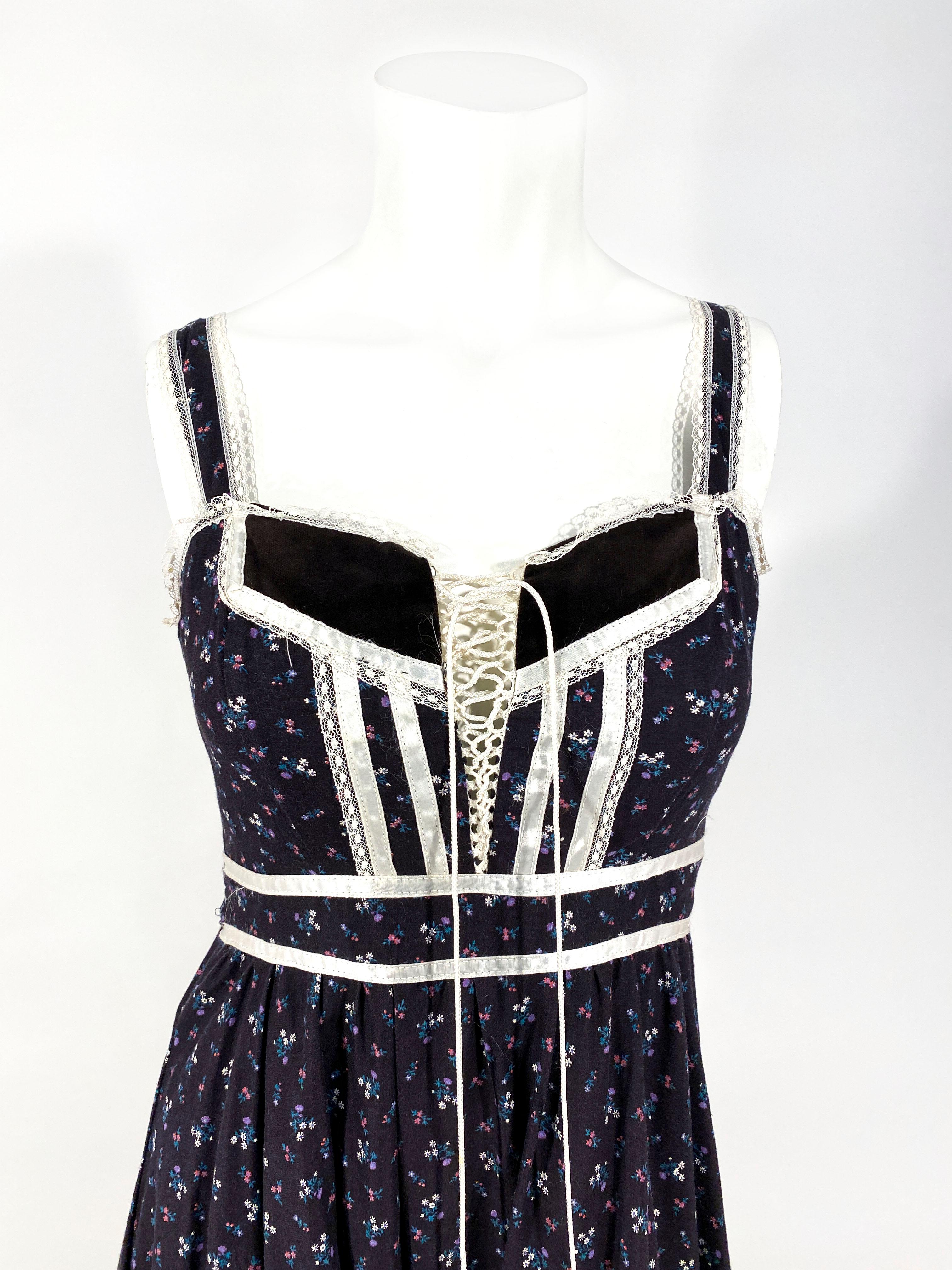 1970s Gunne Sax navy cottage dress featuring a floral calico print, satin and lace trim, velvet lace up bodice, a wide ruffled hem, and an applied sash that ties in the back over the zipper closure. 