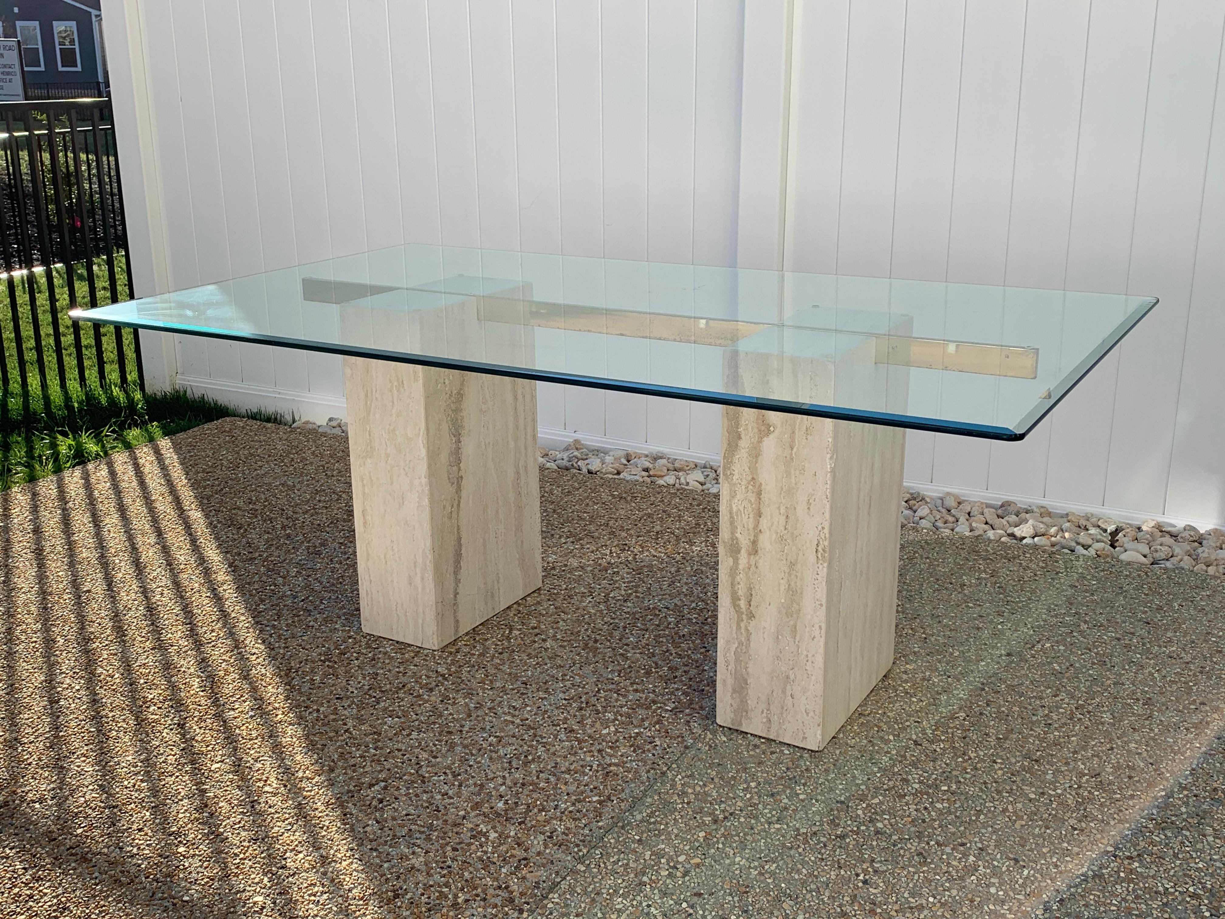 Offered is a stunning, 1970s Italian limestone/travertine dining table by Guy Barker for Ello Furniture Company. The piece has a thick 0.75in tempered beveled glass top, rested upon a single polished brass arm and two limestone/travertine bases.