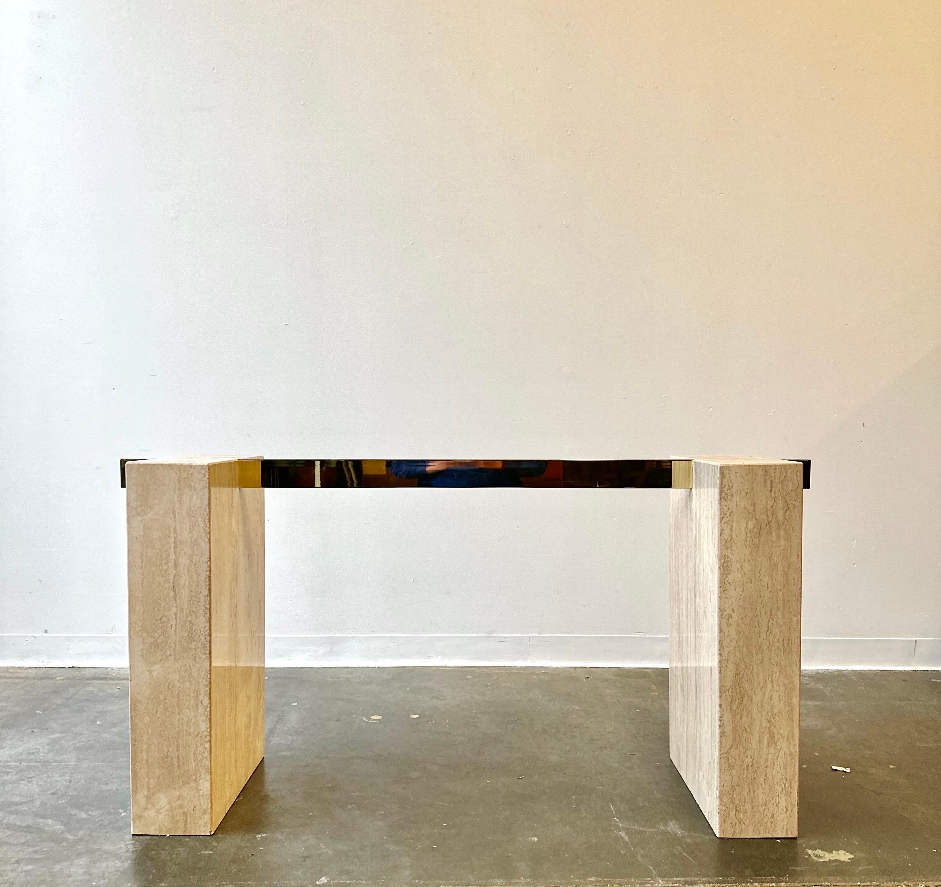 Offered is a stunning, 1970s Italian limestone/travertine dining table by Guy Barker for Ello Furniture Company. The piece has a thick 0.75in tempered beveled glass top, rested upon a single polished brass arm and two limestone/travertine bases.