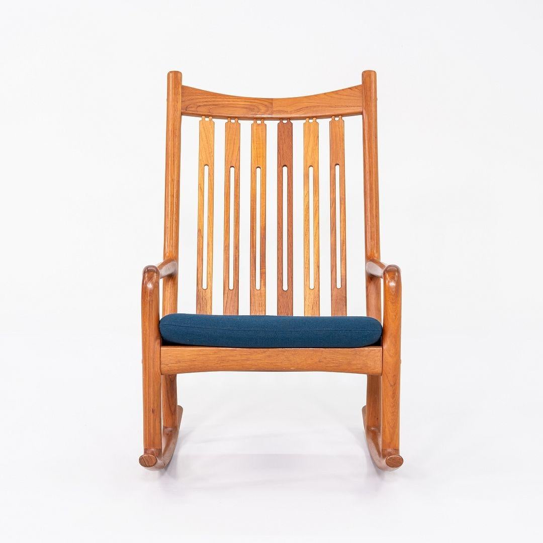 Si tratta di una sedia a dondolo in teak, disegnata originariamente da Hans Olsen per Juul Kristensen. Questo particolare esemplare risale agli anni '70. La struttura della sedia è in teak massiccio e presenta un cuscino imbottito in tessuto blu.