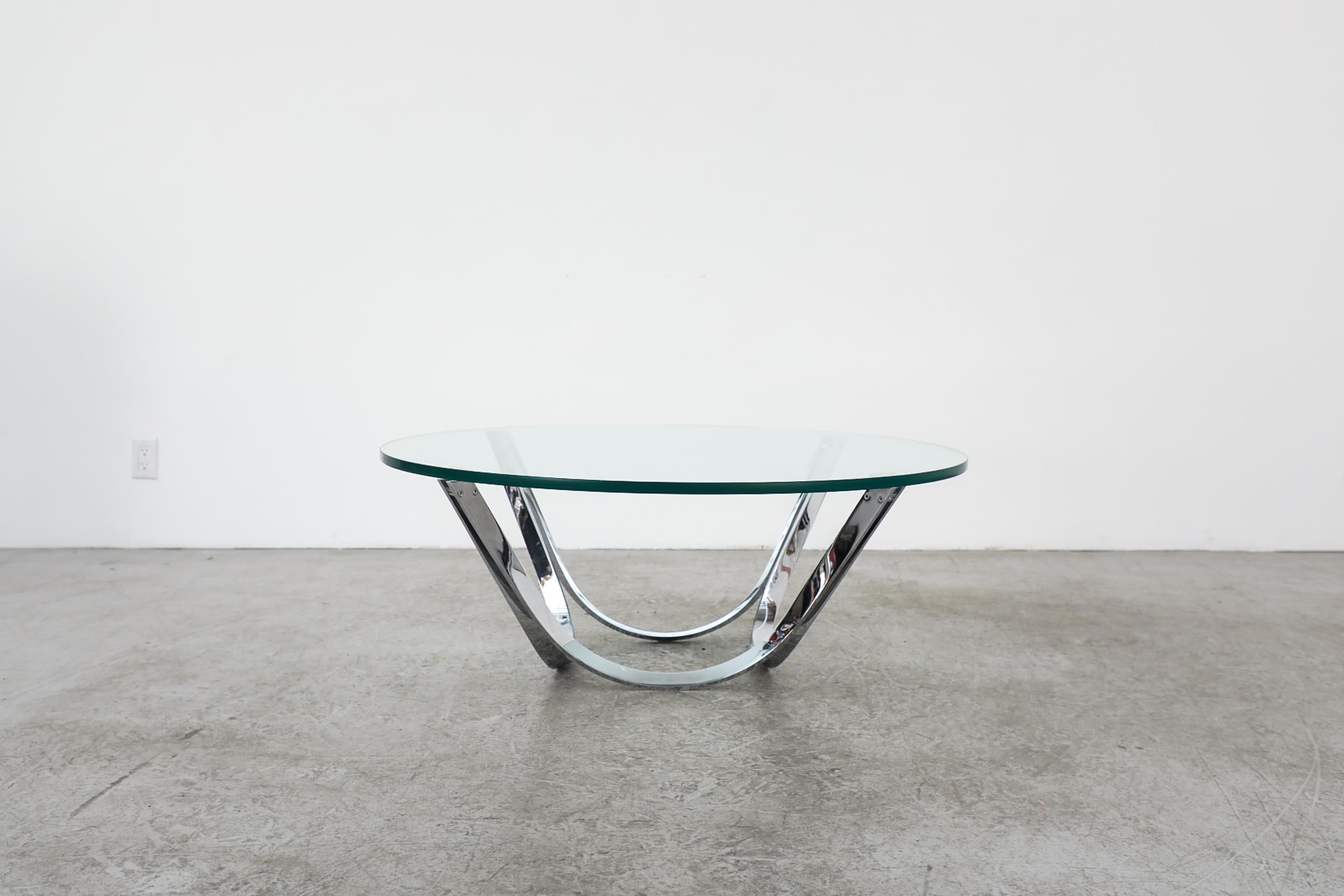 This Mod 1970's coffee table has a striking, heavy chrome frame with curved legs and a thick plate glass top. It's in original condition with visible wear, including some scratches and chips on the glass. Wear is consistent with its age and use.