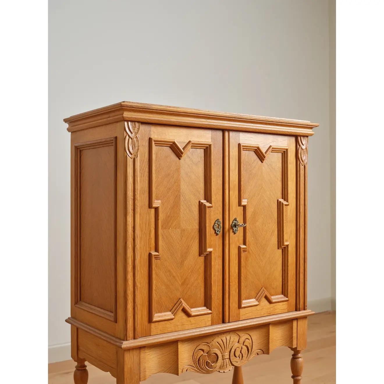 Dry bar in quarter-sawn oak; carved with stylized leaves at the trim and a scrolled fan or sheaf of wheat motif at the lower drawer front, featuring Gothic-inspired recessed panel molding at the doors. Numbered 3865 on back panel. Designed by