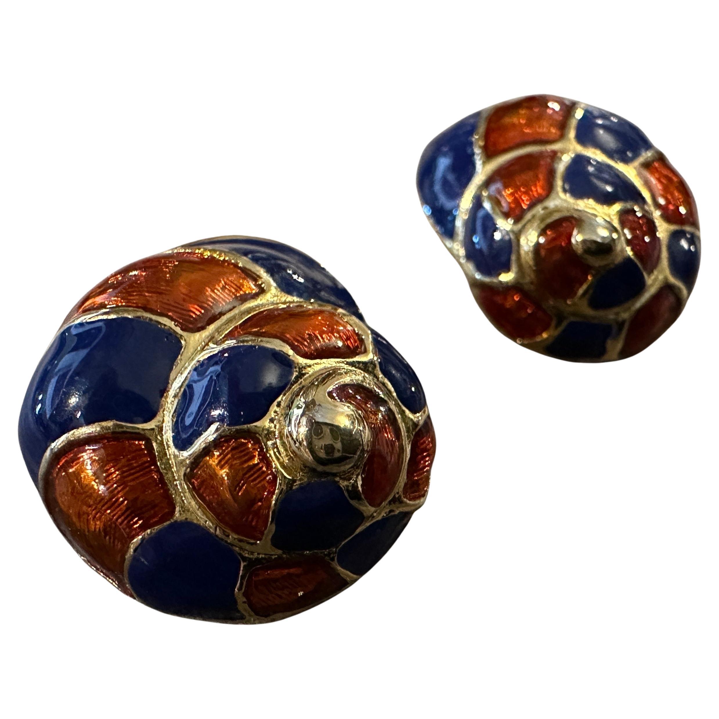 Dieses Paar Vintage-Ohrringe besticht durch ein charmantes Muschelmuster, das mit leuchtender Emaille in tiefblauen und warmen rot-orangen Tönen verziert ist. Das verschlungene Muster ist mit goldfarbenen Akzenten umrandet, die den schneckenartigen