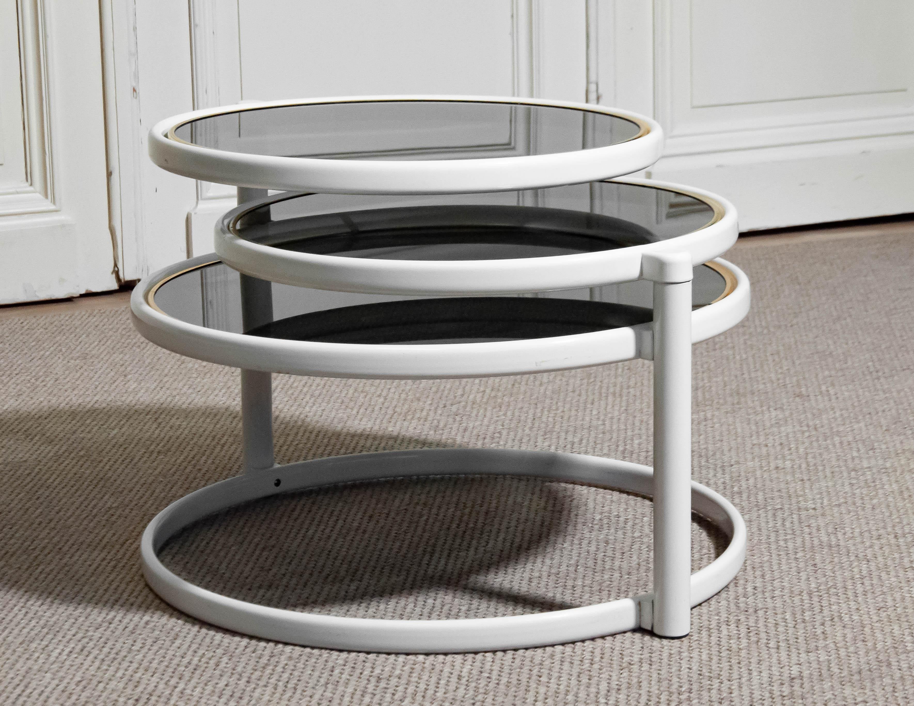 Si tratta di un tavolino da caffè degli anni '70 nello stile di Milo Baughman, caratterizzato da un design elegante e moderno. Il tavolo ha una struttura in metallo bianco con una base circolare centrale che sostiene tre piani d'appoggio rotondi.