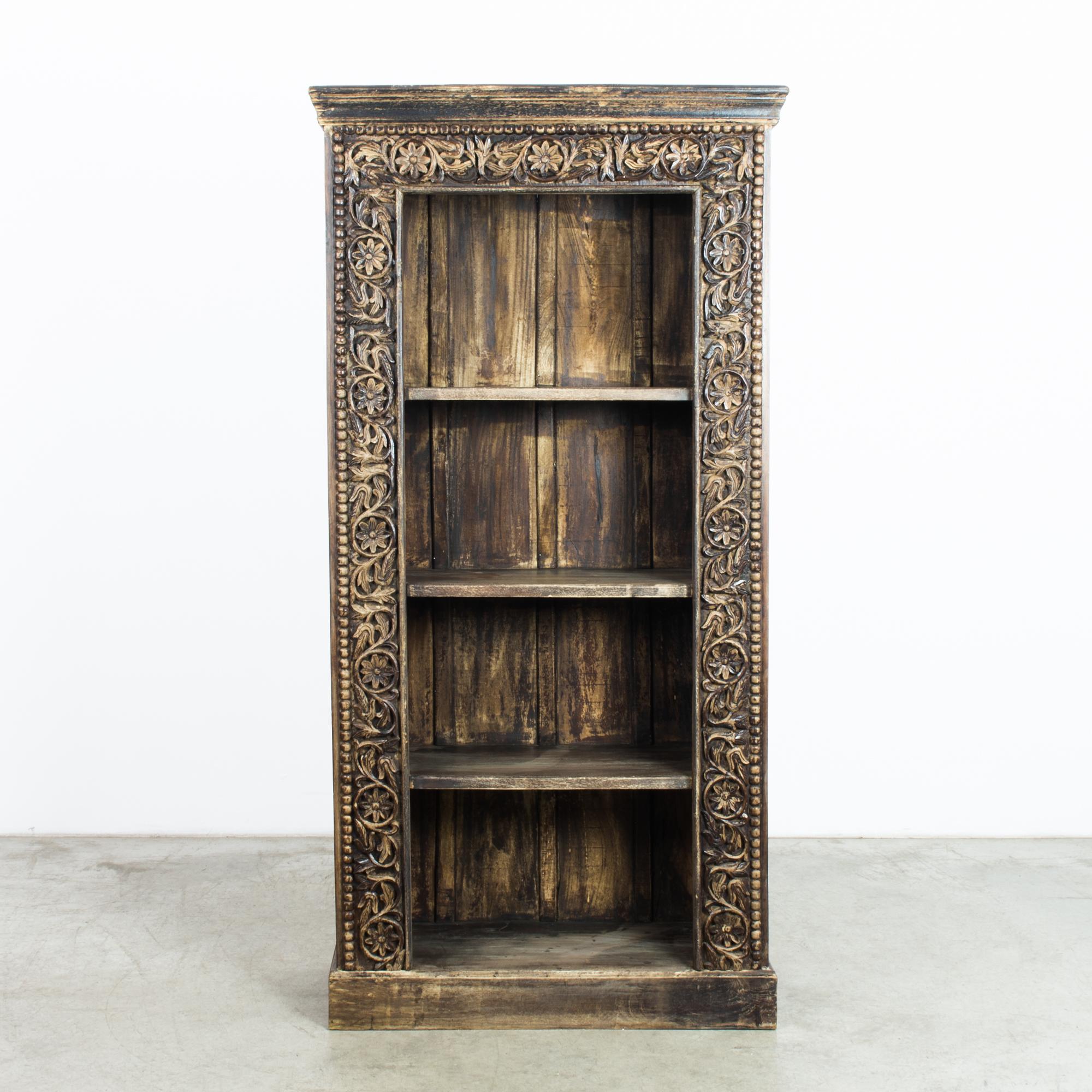 This wooden bookshelf with crown molding was made in India, circa 1970. The ornate carvings with ringed flowers and leaves once framed a collection of books resting on the four sturdy shelves. The finish has mellowed with age to display a timeworn