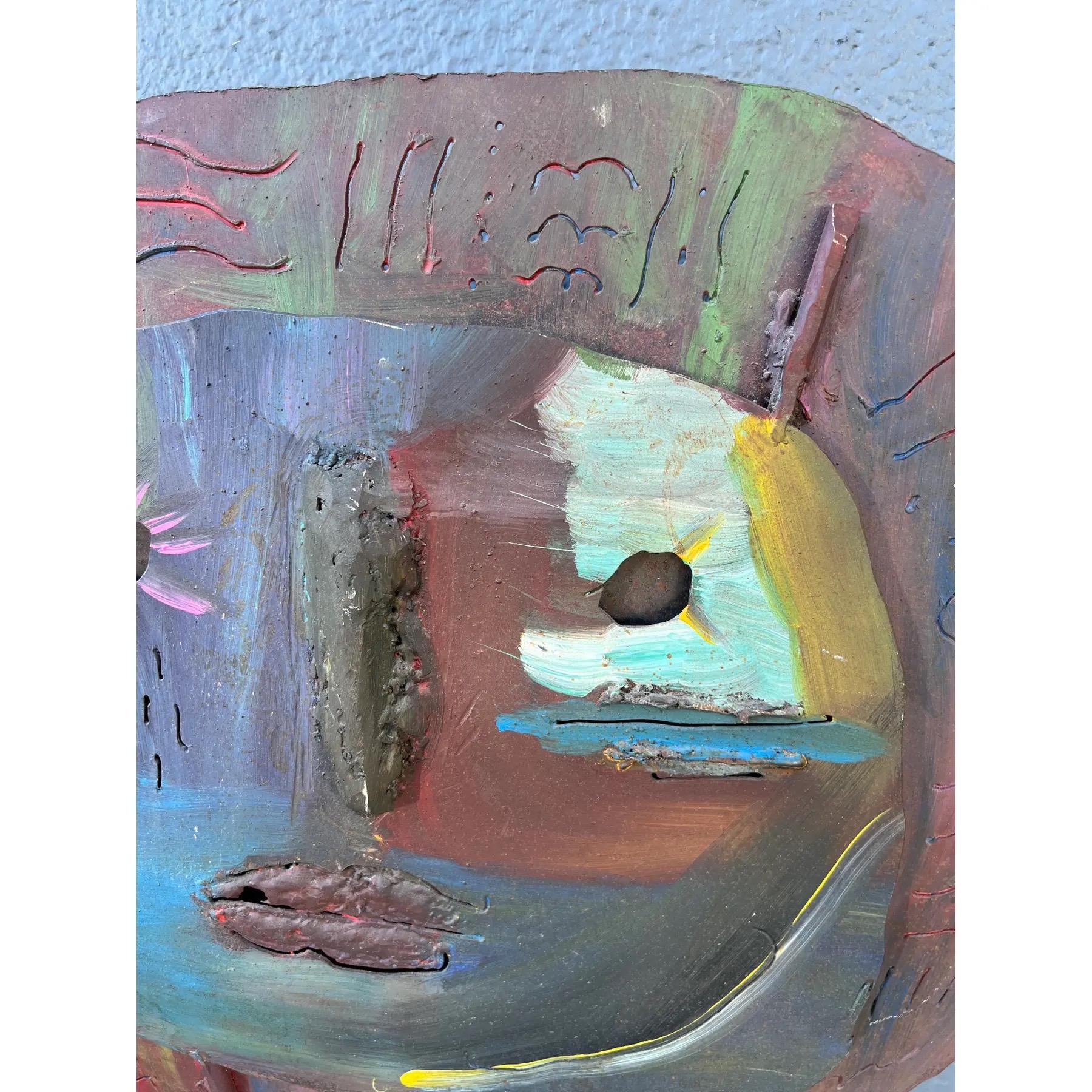 A modernist sculpture in iron. Depicts a round face with simple raised and pierced features, decorated in a vibrant color palette. Artist is unknown.