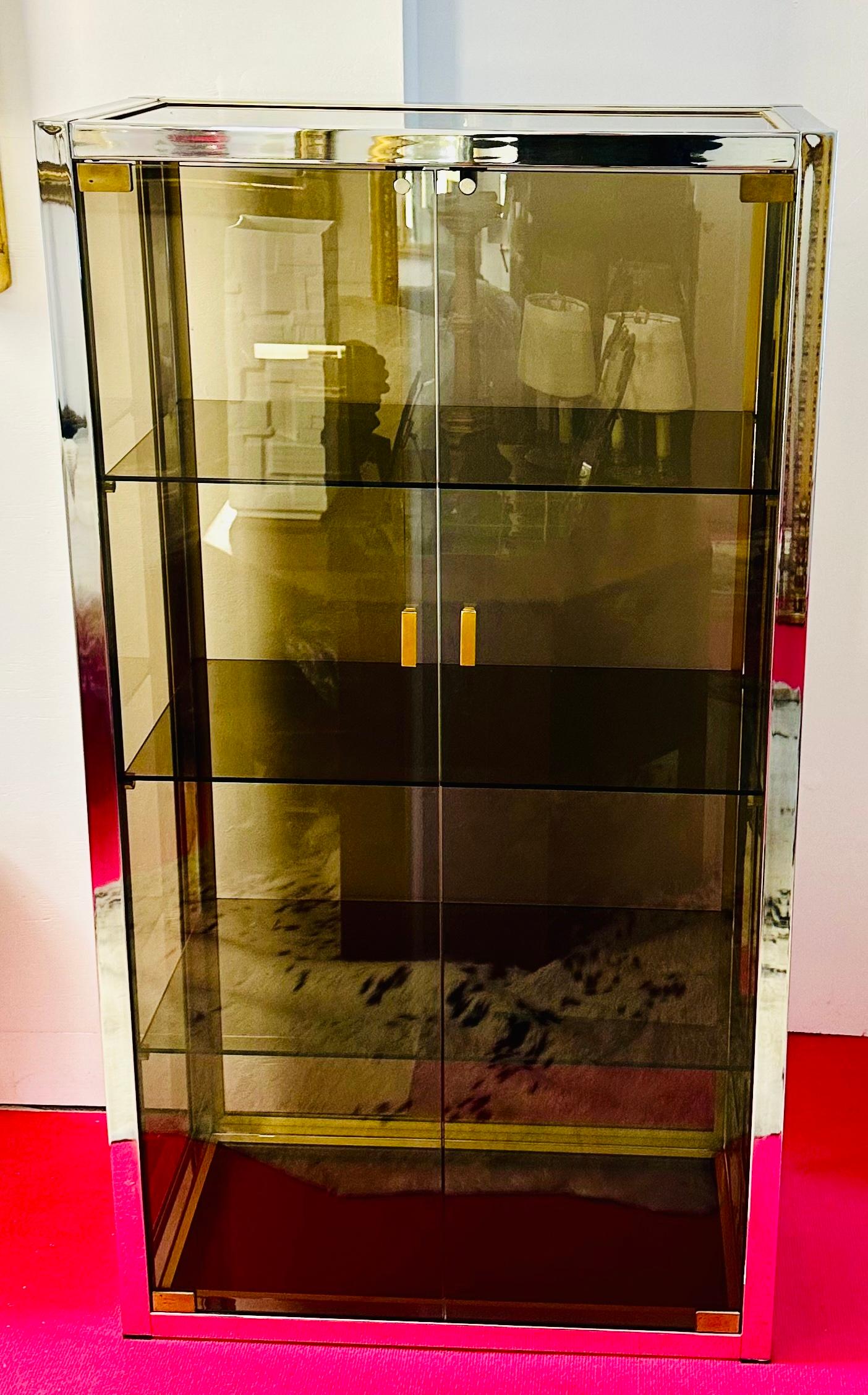 An Italian smoked glass, chromed-silver and chromed-brass display or drinks cabinet designed by Renato Zevi during the 1970s.  The Hollywood Regency style cabinet has three fixed smoked-glass display shelves and doors with brass door handle-pulls. 