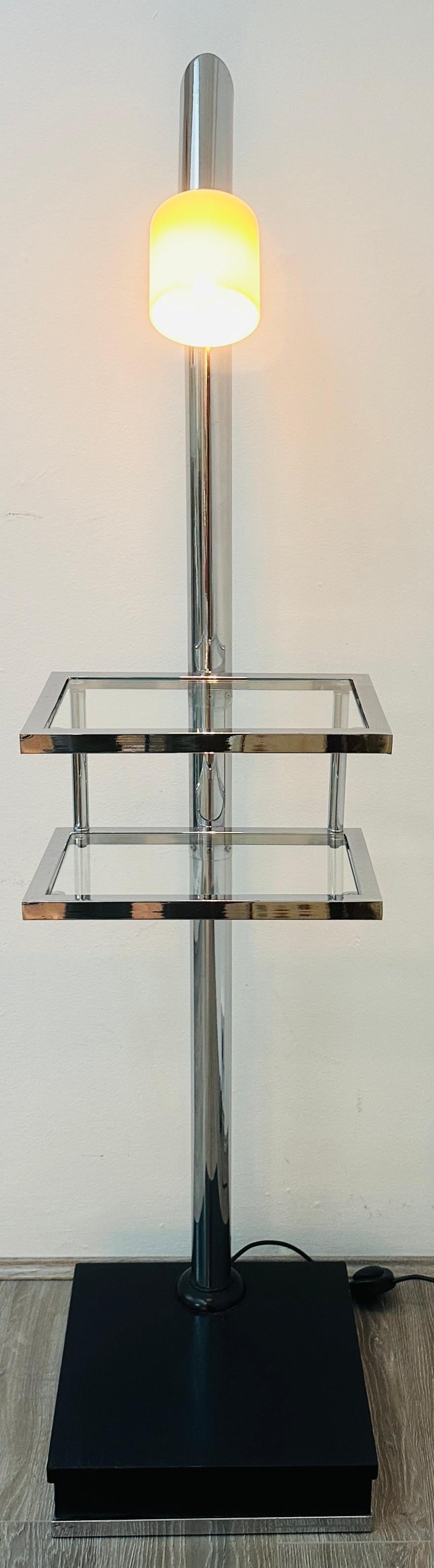 Lampada da terra italiana degli anni '70 in cromo lucido che incorpora due piccoli ripiani con inserti in vetro temperato trasparente. La colonna portante in tubolare di metallo cromato che sostiene i due ripiani paralleli è sostenuta da una base