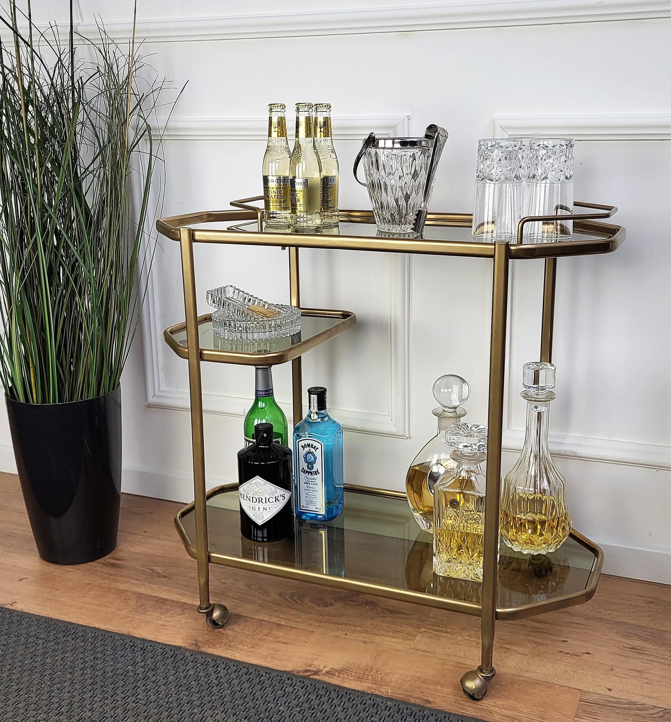 Beautiful and stylish 1960s Hollywood Regency Italian octagonal two-tier metal brass color and smoked glass bar cart with removable top tray and small half-way side shelf. Very good condition of both the brass finish and the glass trays.

A great