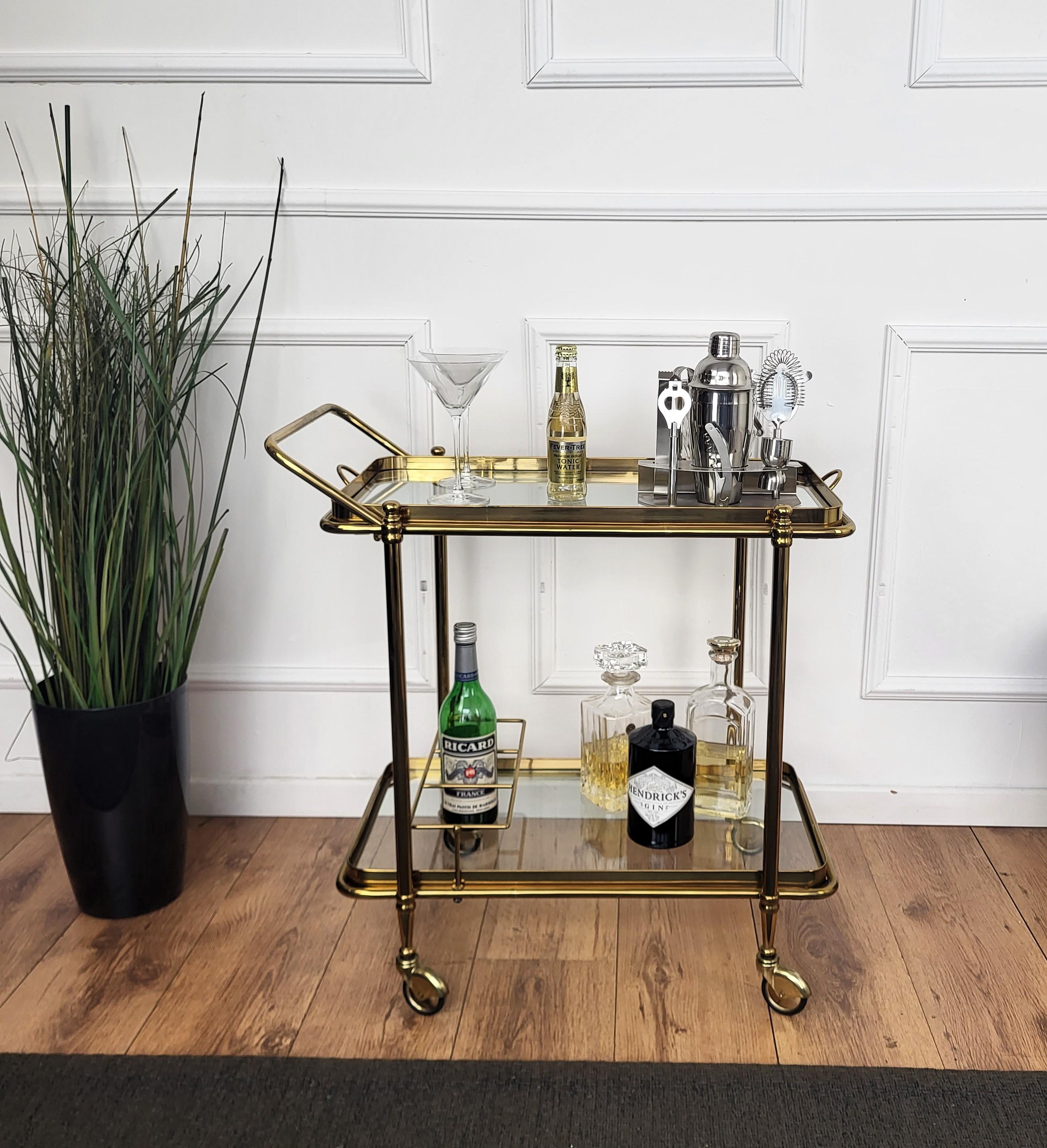 Beautiful and stylish 1960s Hollywood Regency Italian two-tier shiny gilt brass and glass bar cart with removable top tray and squared 3 bottle holder side shelf. Very good condition of both the brass finish and the glass trays.

A great piece that