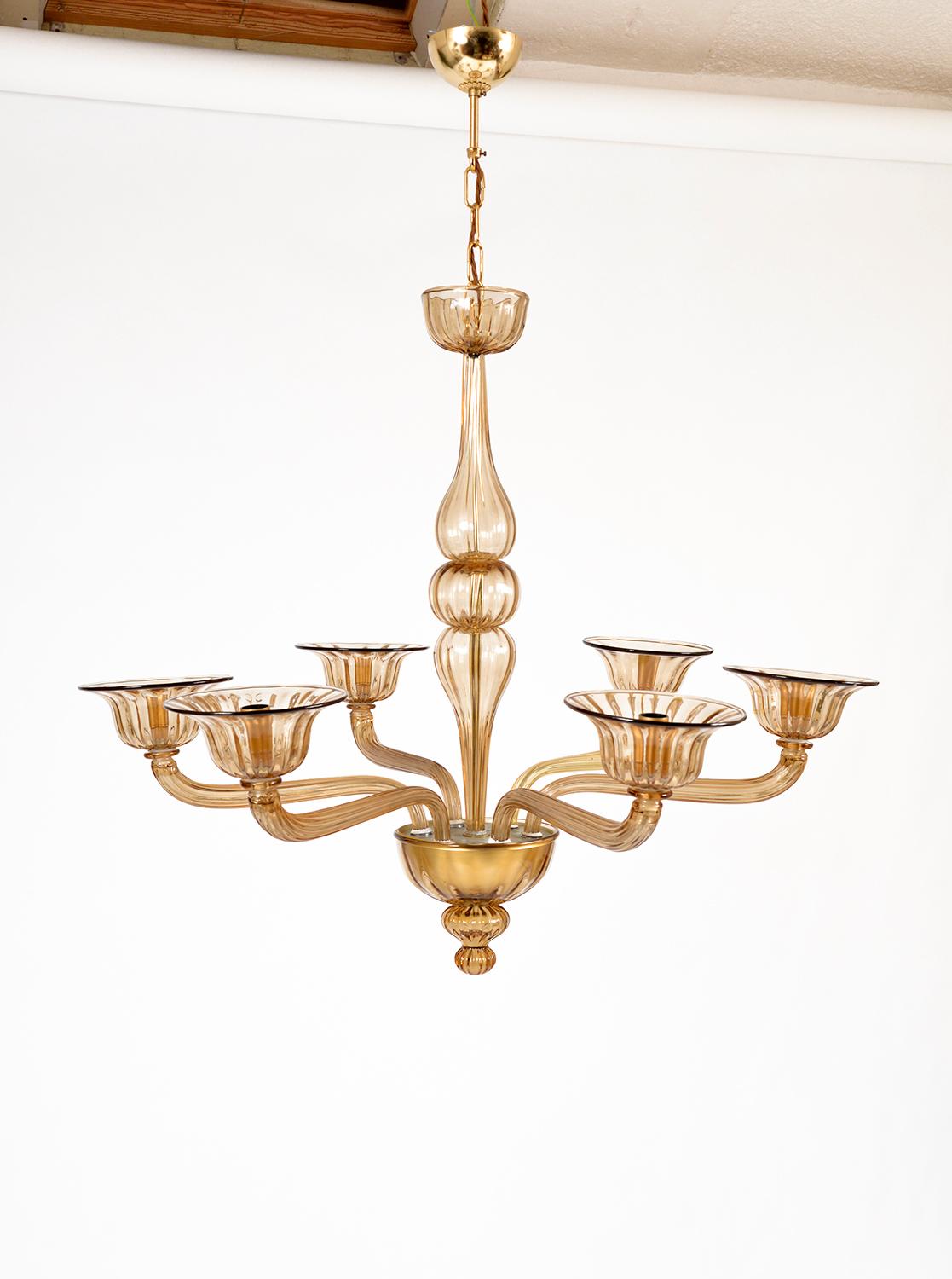 A classic six-arm Venetian Murano chandelier in hand-blown amber coloured glass. The scroll arms with ribbed decoration, hold six flared bobeche where the light bulbs are nested, culminating in a bowl-shaped gold base with a teardrop finial.
This