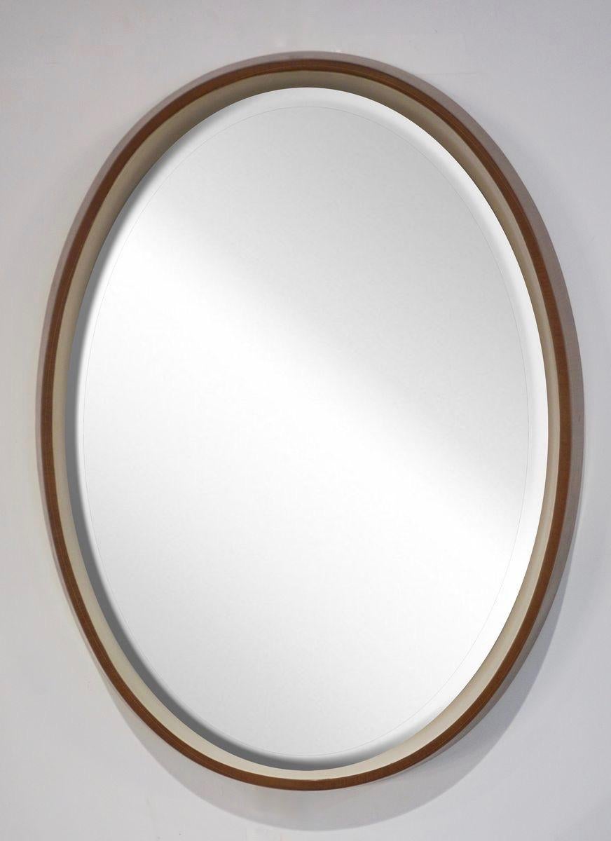 An Italian Mid-Century Modern design mirror in an interesting oval shape, original quality mirror with beveled edge, backlit and raised in a white painted wood interior to enhance the light source and maximize the illumination, framed by a