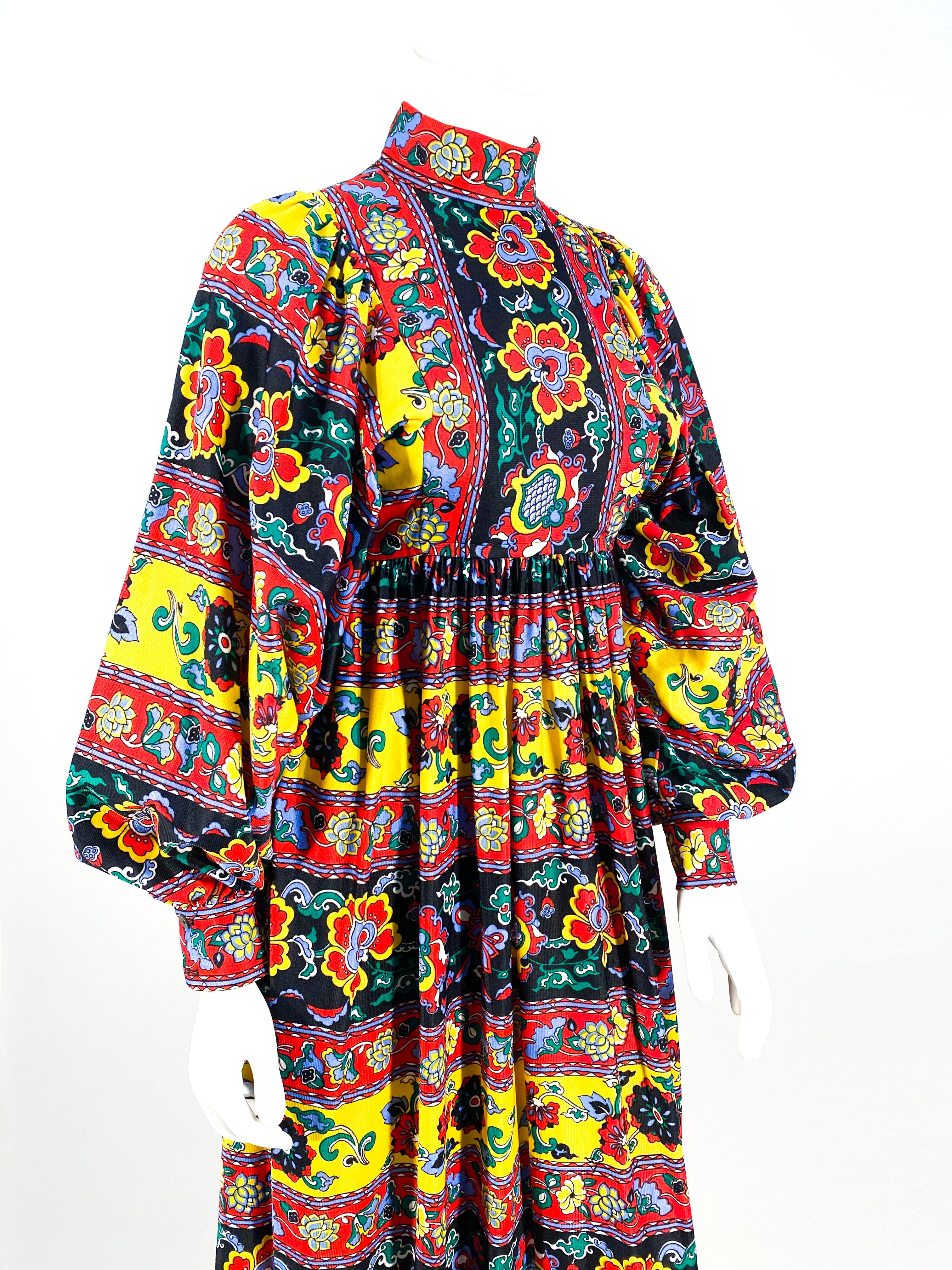 1970s stripped paisley printed knit polyester jersey peasant dress in jewel-tones of, navy, red, yellow, green, red, and blues. The silhouette of the dress features a full length skirt, a high-neck collar, and enlarged full cuffed sleeves. The back