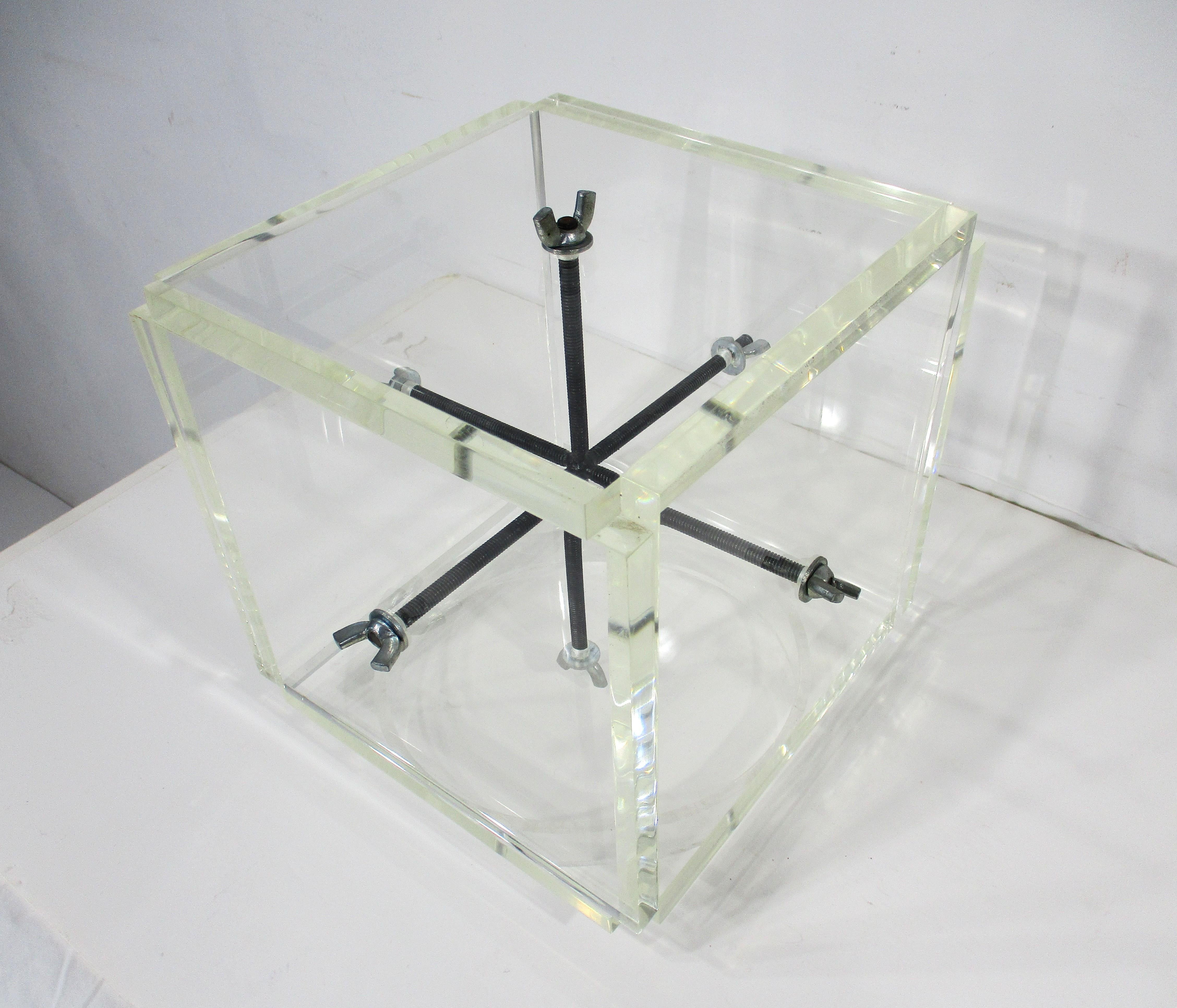 Lucite spessa e lastricata in un cubo tenuto insieme da grandi viti e dadi ad alette che conferiscono al pezzo un aspetto da puzzle industriale. Creato dall'artista, scultore, designer e architetto John Petersen che è stato uno dei fondatori del