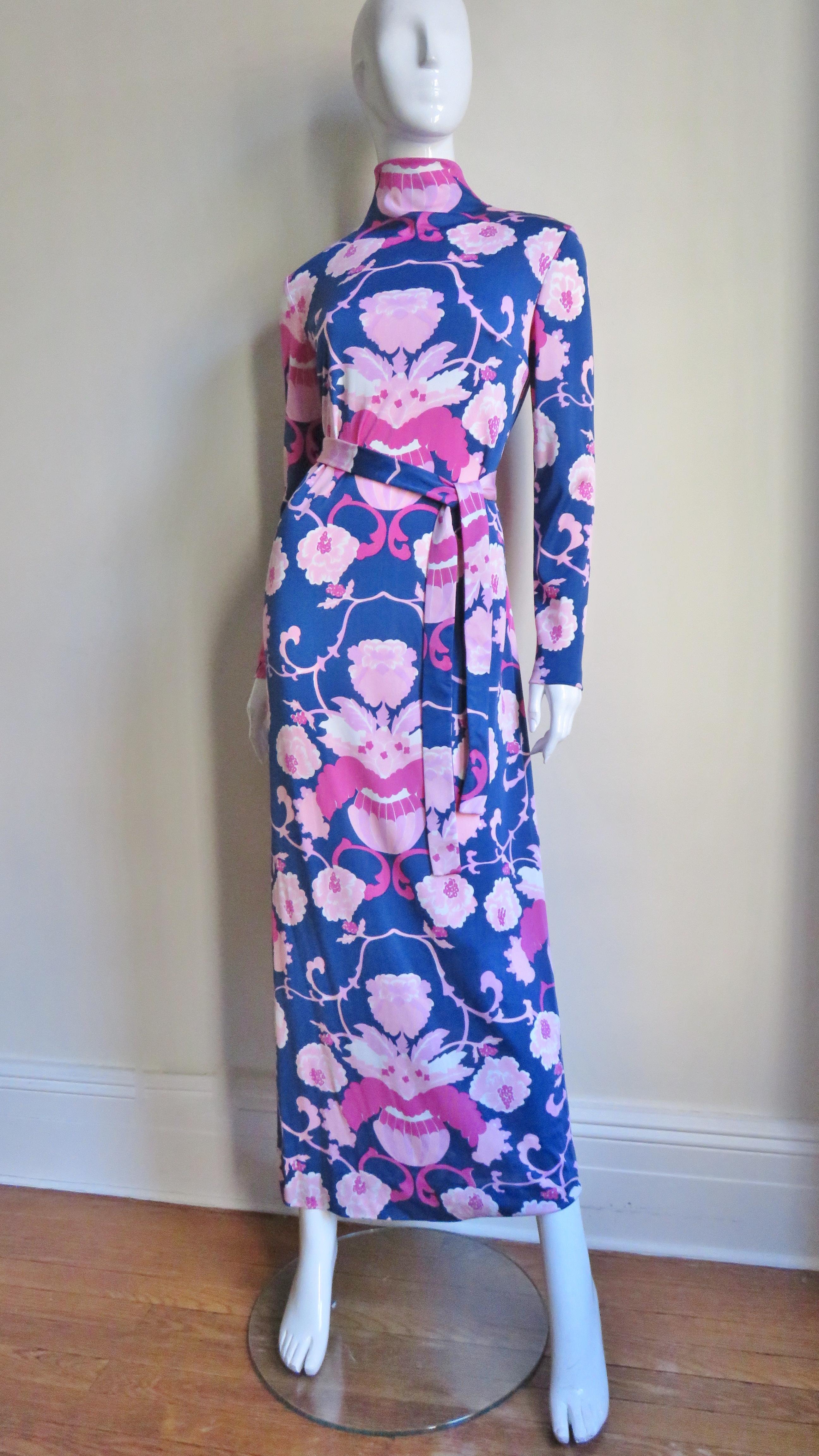 A 3 piece jersey and silk set in a pattern of flowers in various shades of pink on a blue background. The long sleeve sheath maxi dress comes with an optional matching tie belt and a matching full semi sheer silk over skirt in the same pattern.  The