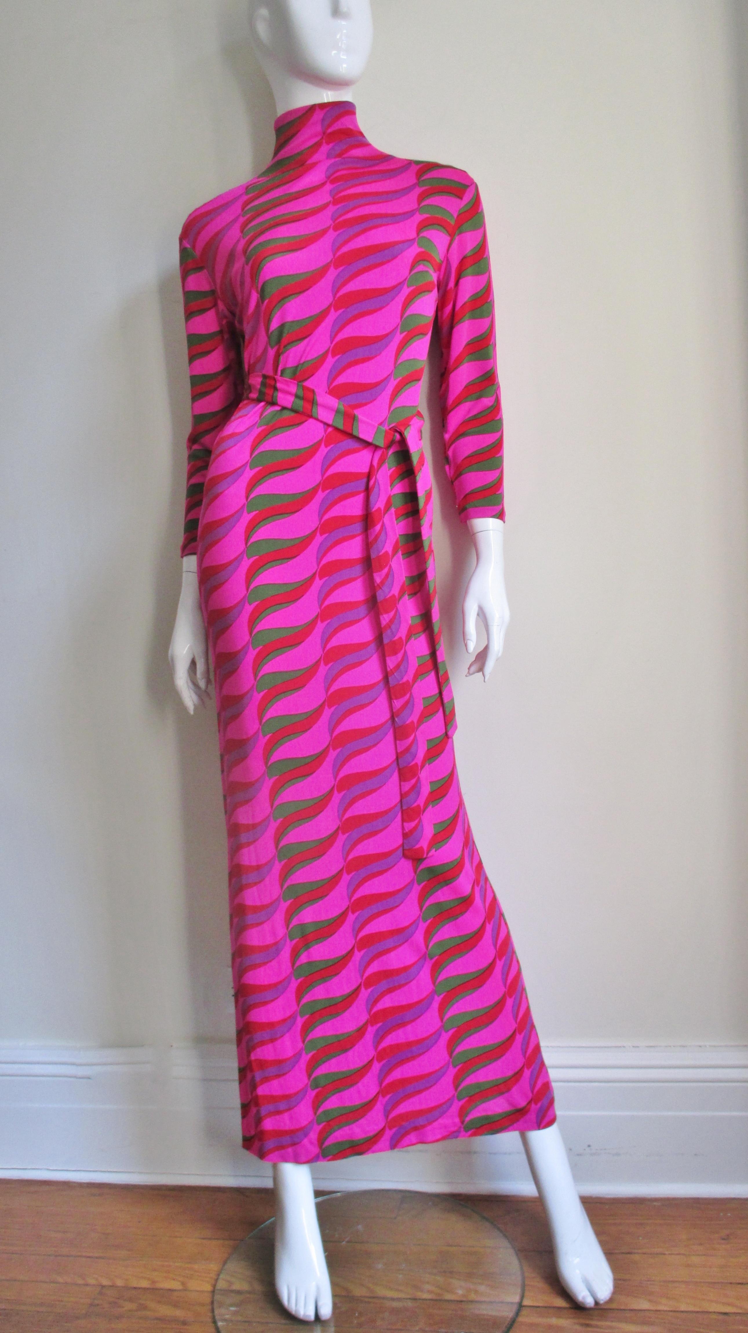 A silk knit long dress from La Mendola in a green, purple and red abstract pattern on a vibrant pink background.  It is a simple full length sheath with a stand up collar and 3/4 length sleeves.  The back closes with a row of self covered buttons