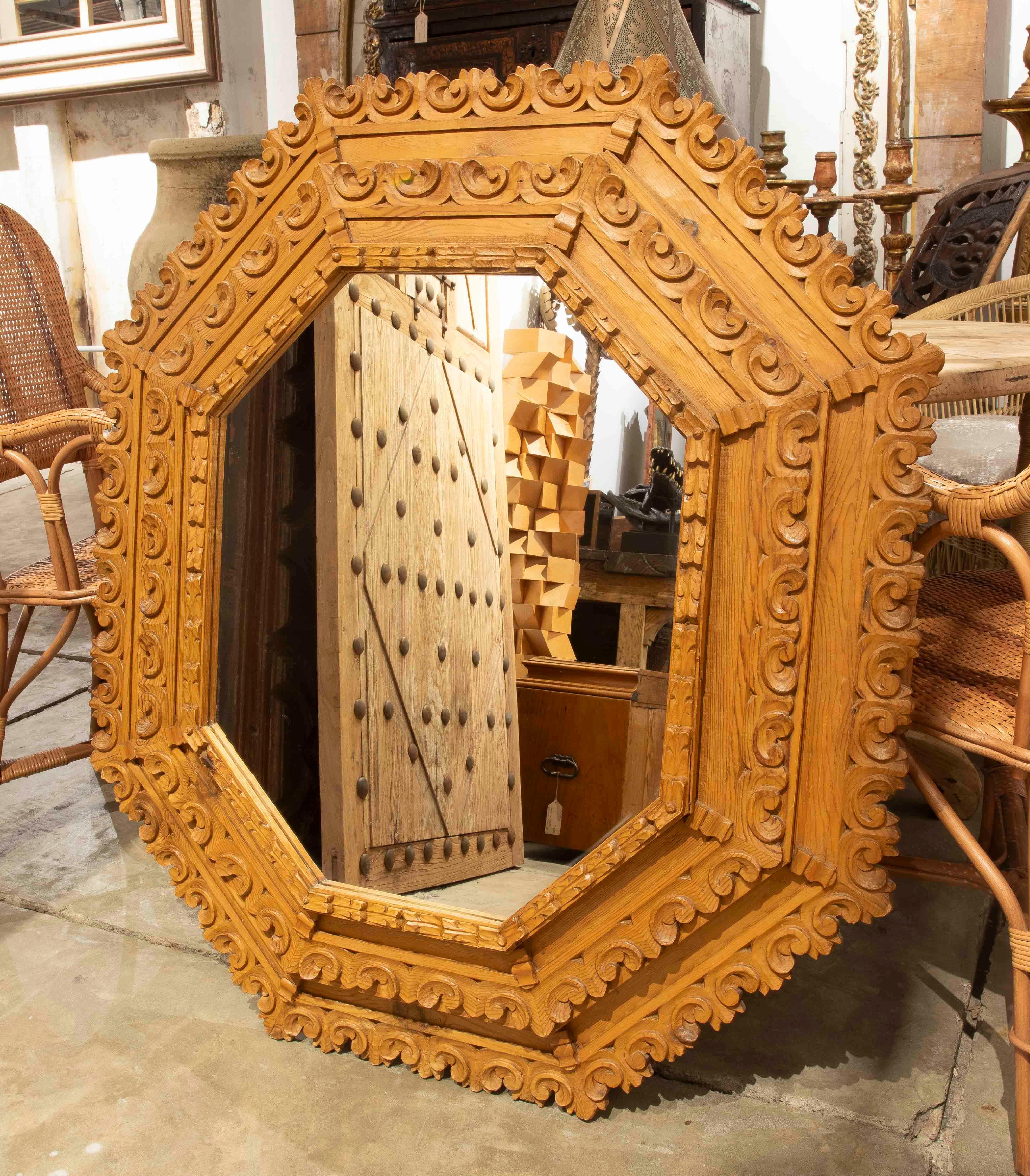 Imponente espejo octogonal de gran formato con marco artesanal de madera natural. El diseño ornamentado, con volutas, hojas y molduras escalonadas, recuerda al estilo barroco, destacando por su simetría y profundidad.

Una pieza decorativa de gran