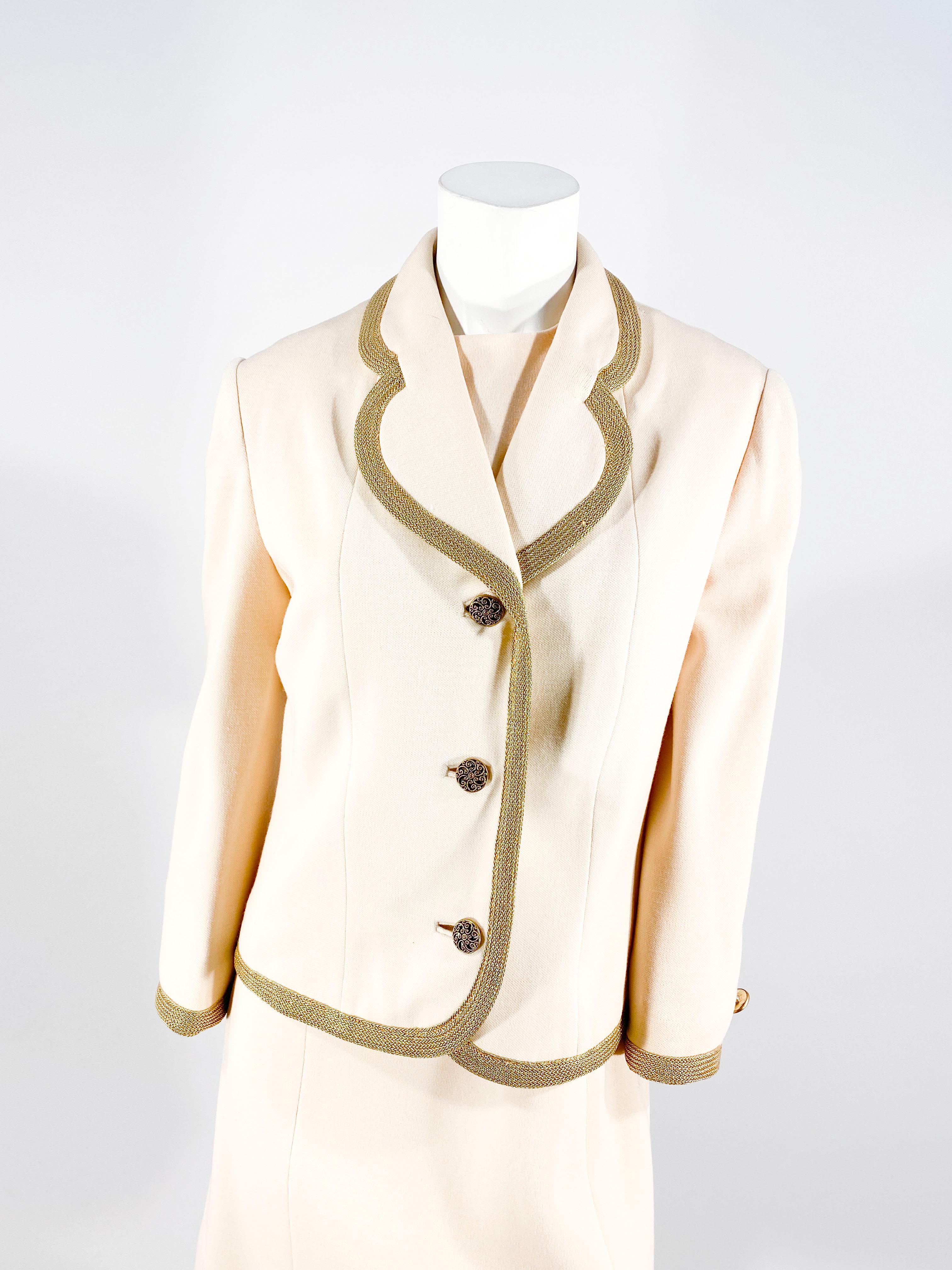 1970s Lilly Ann Ivory-colored wool knit suit with metallic brass trim along the collar and coat edges. The knee length shift dress is sleeveless and entirely lined with a back zip closure. The jacket has designer buttons along the face and the