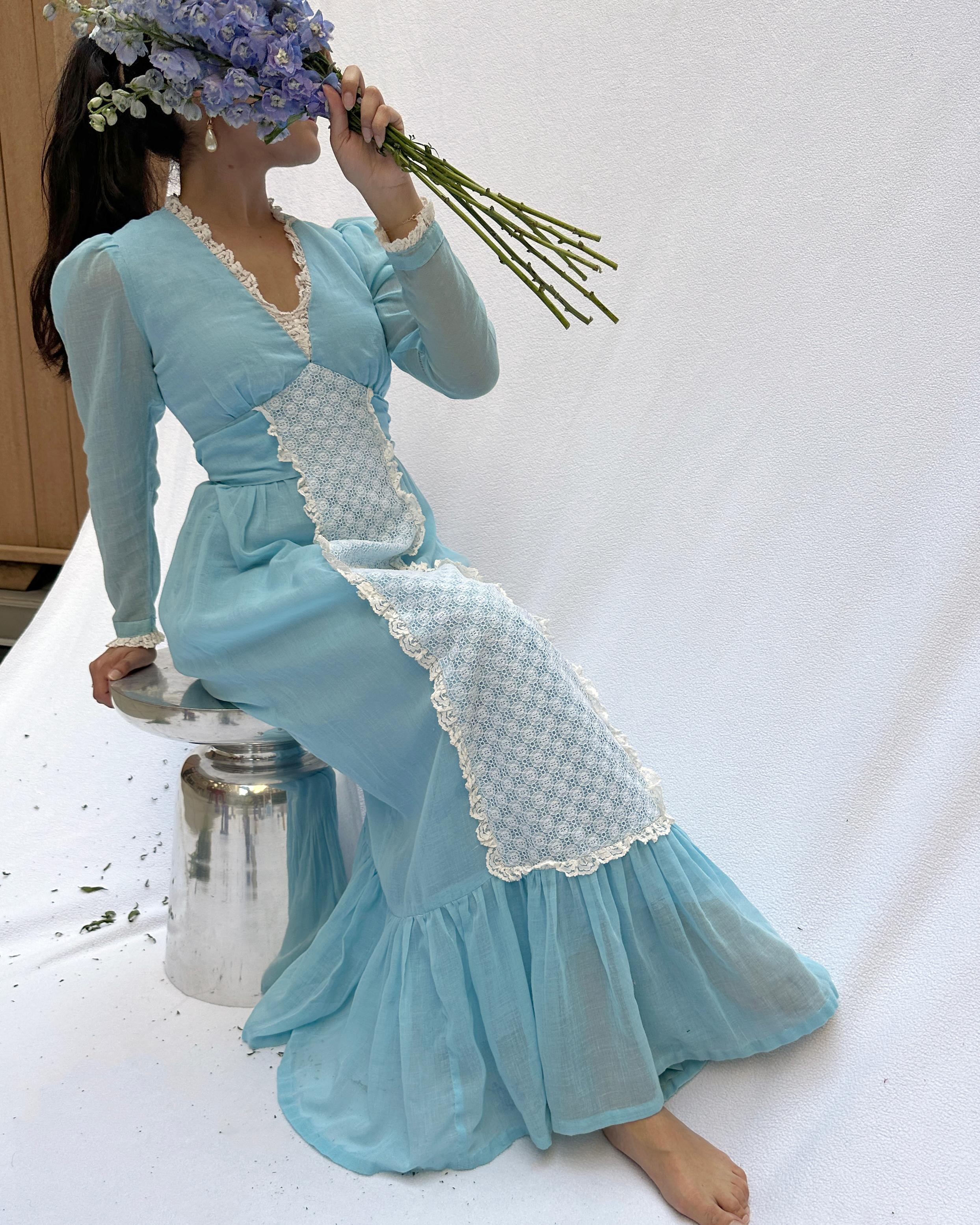 This vintage Gunne Sax style dress is so 1970s-does-Victorian. The blue color is just a dream! It features lace overlay down the front, a flounce at the hem, and lace trim throughout. The empire waist features a ribbon belt that ties in the back in