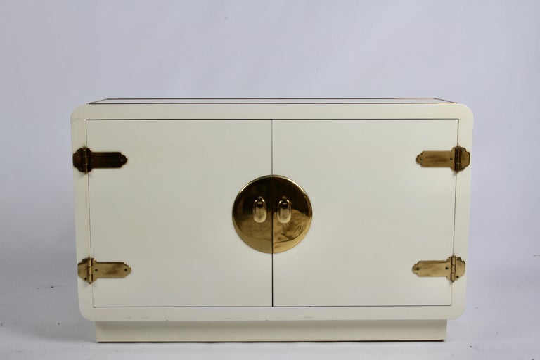 1970s Mastercraft designed Hollywood Regency Asian inspired creme lacquered cabinet, TV stand or entry console with Asian inspired brass hardware and inlaid trim details. This two door cabinet has one adjustable interior shelf, and its narrow stance