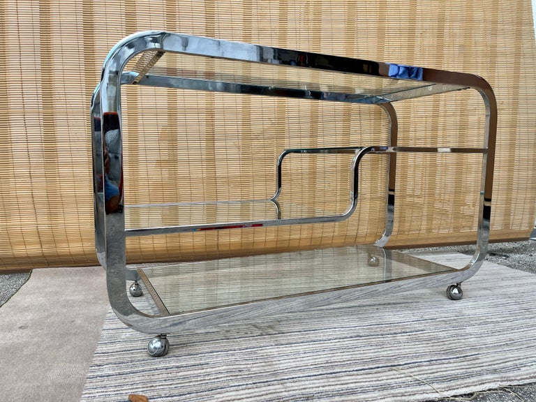 Vintage Mid-Century Modern Hollywood Regency Chromed rolling dry bar cart in the Milo Baughman Style. Circa 1970s.
Features a very stylish bent chromed steel frame with four tempered removable clear glass shelves to accommodate bottles, glassware,