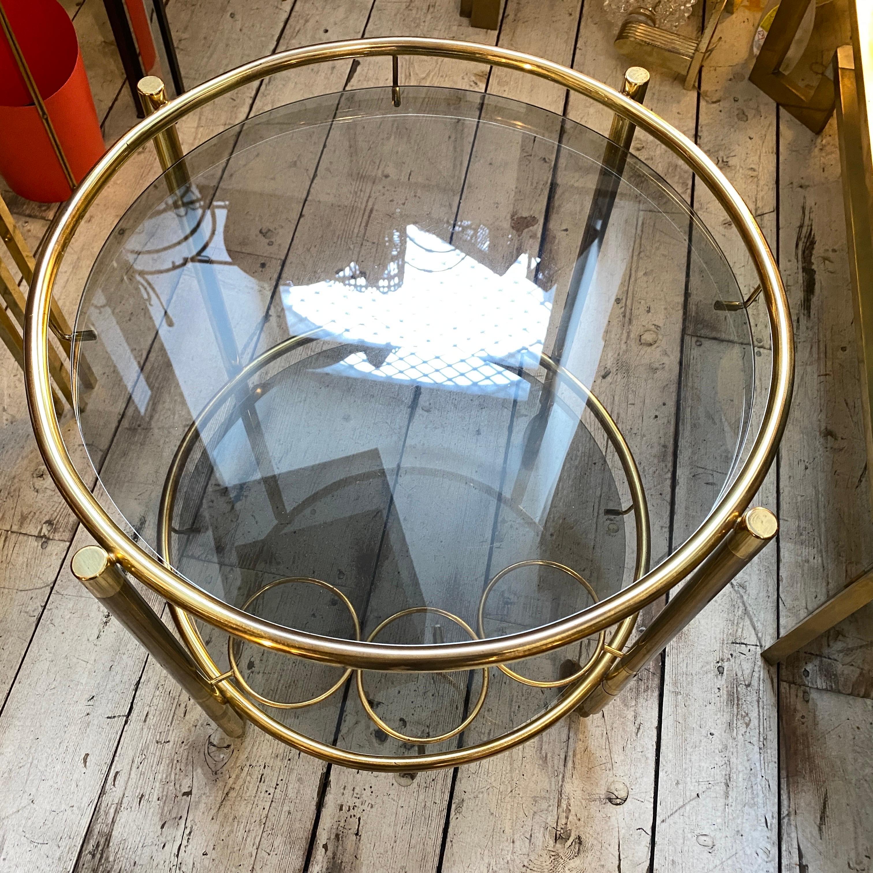 A very good conditions, brass and smoked glass italian bar cart, it has been made in the Seventies and the original brass patina gives it a stunning vintage look. This round bar cart is a stylish and functional piece of furniture that captures the