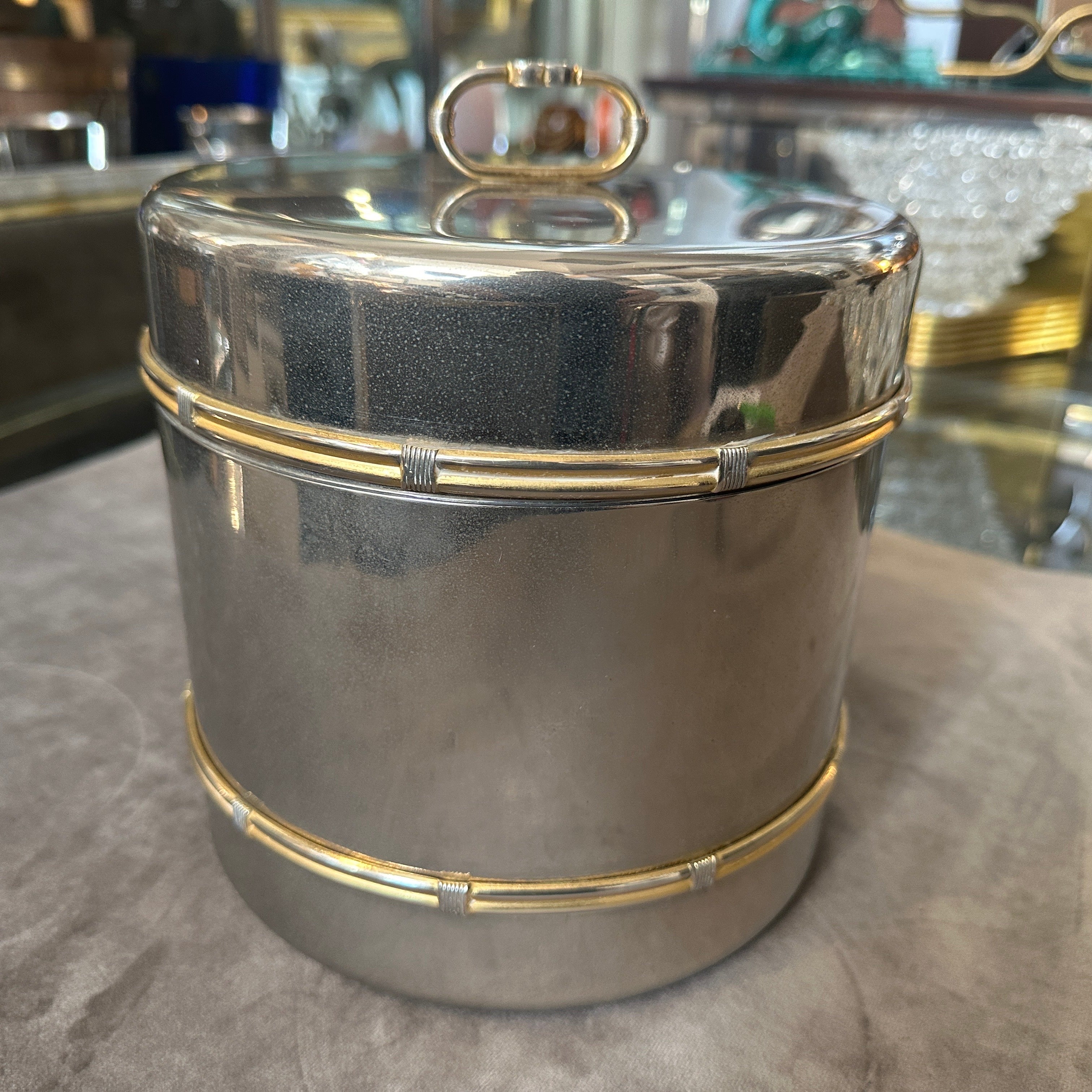 This 1970s Gucci ice bucket exemplifies the opulence and craftsmanship synonymous with the brand during this era. The primary body and lid of the ice bucket are crafted from silver plate, giving it a sleek and reflective finish.
The bucket features