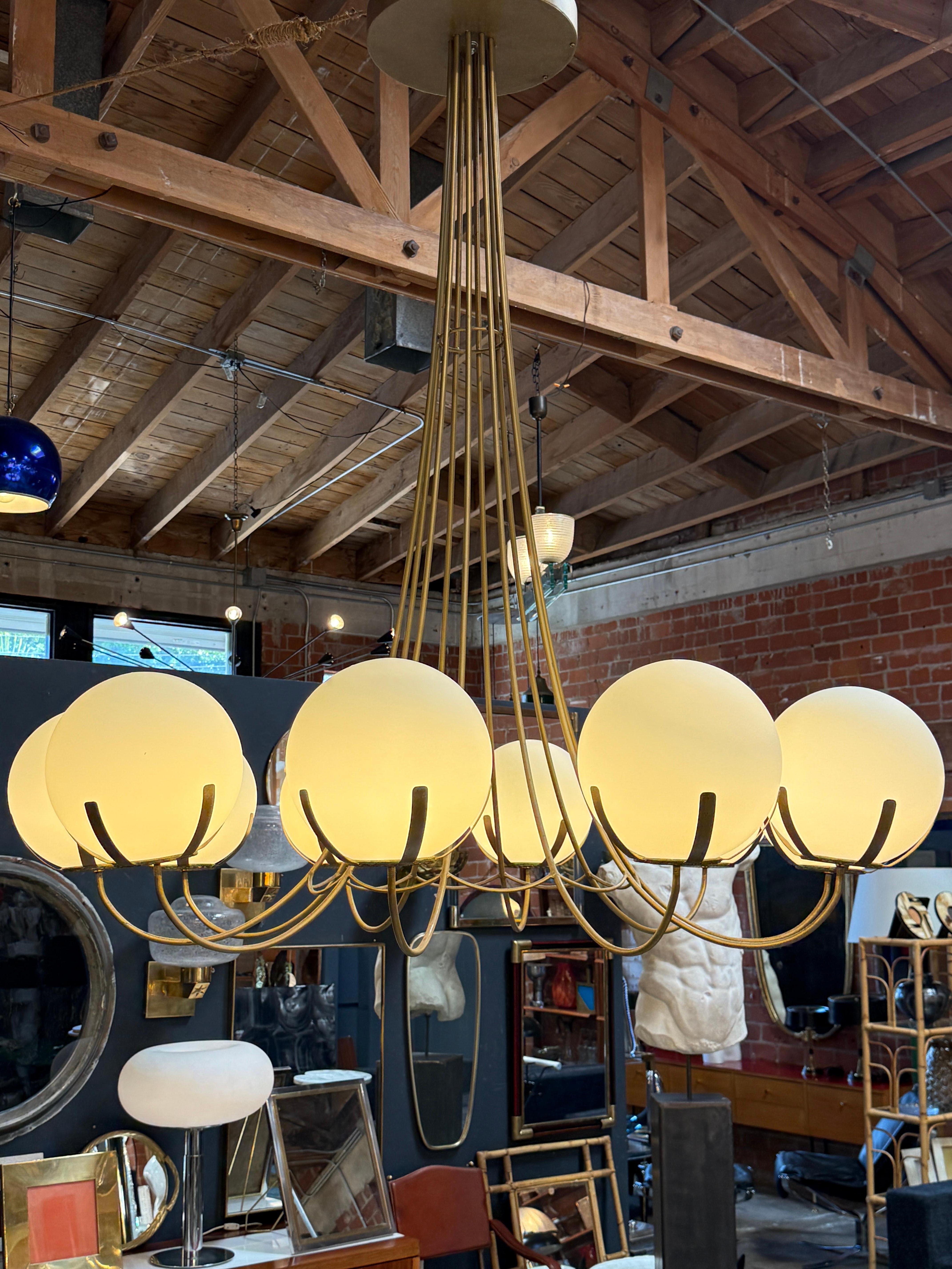 This striking oversized Italian chandelier, crafted in the 1970s, recalls the graceful geometry of a carousel at rest—perfectly still, yet full of quiet energy. Its solid brass frame supports ten frosted glass spheres that emit a soft, diffused