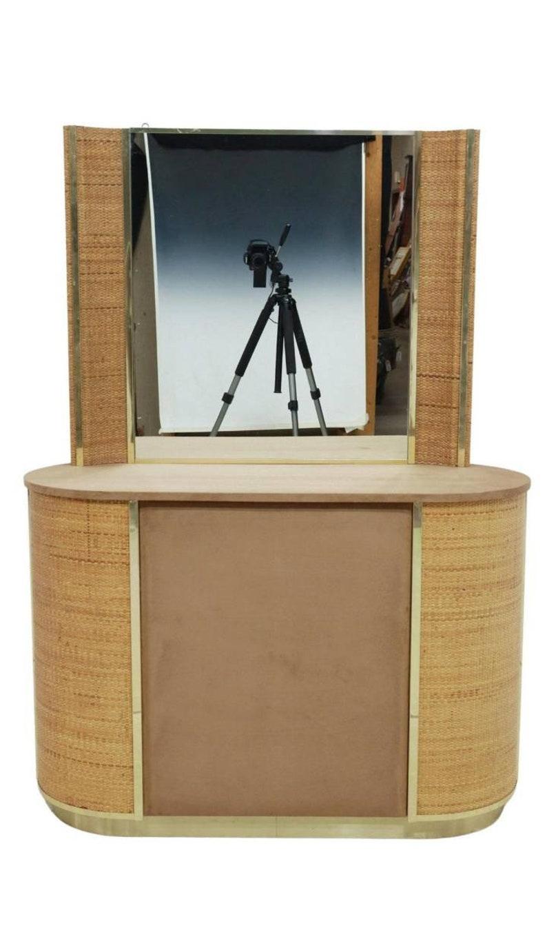 Une console d'entrée vintage post-moderne / coiffeuse et miroir, vers les années 1970, ayant une plaque de miroir plat incurvée en rotin tressé et encadrée de laiton, au-dessus d'une table de forme ovale avec un dessus en daim, des panneaux en rotin