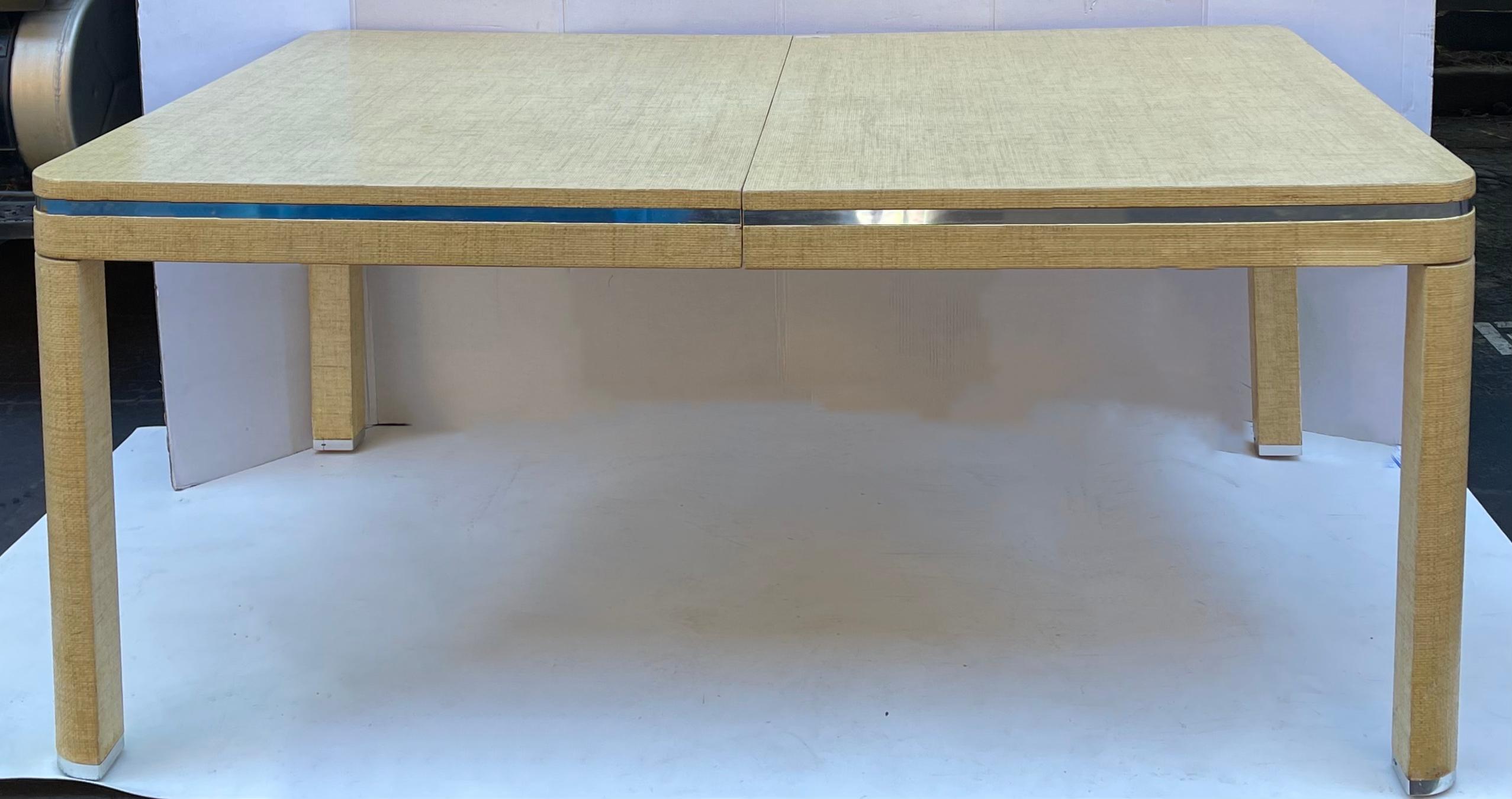 Love this! This is a modern Karl Springer style grasscloth wrapped dining table with two leaves. It has clean, modern styling with chrome capped feet and trim. Each leaf adds 18” to the overall width. The two leaves are lighter than the table. I