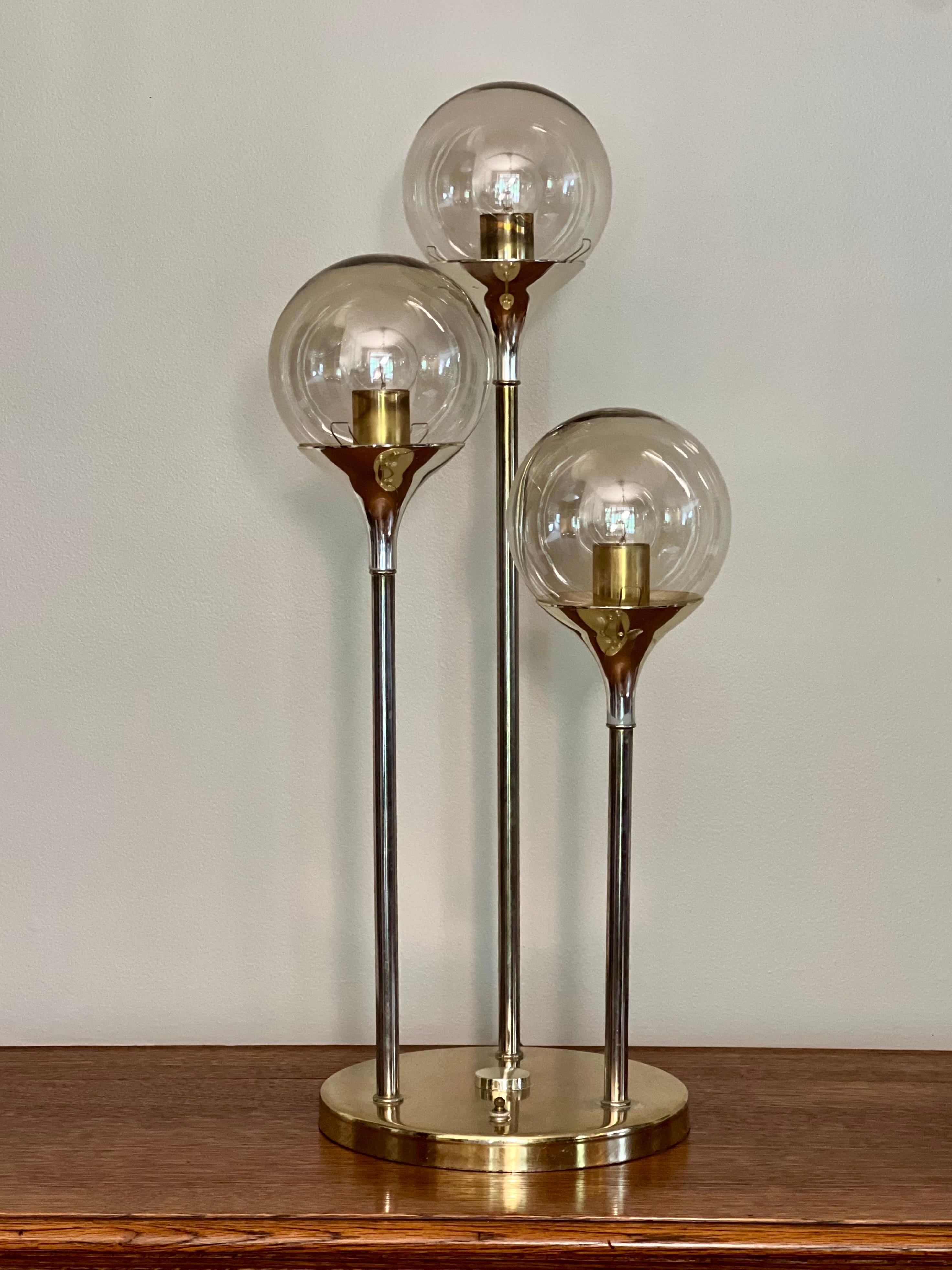 1970s atomic Space Age style three globe table lamp. It features lightly smoked glass globes at tapered lengths, a brass finish and a switch on the base with three settings. Globes are held in place by tension clips making it easy to change bulbs