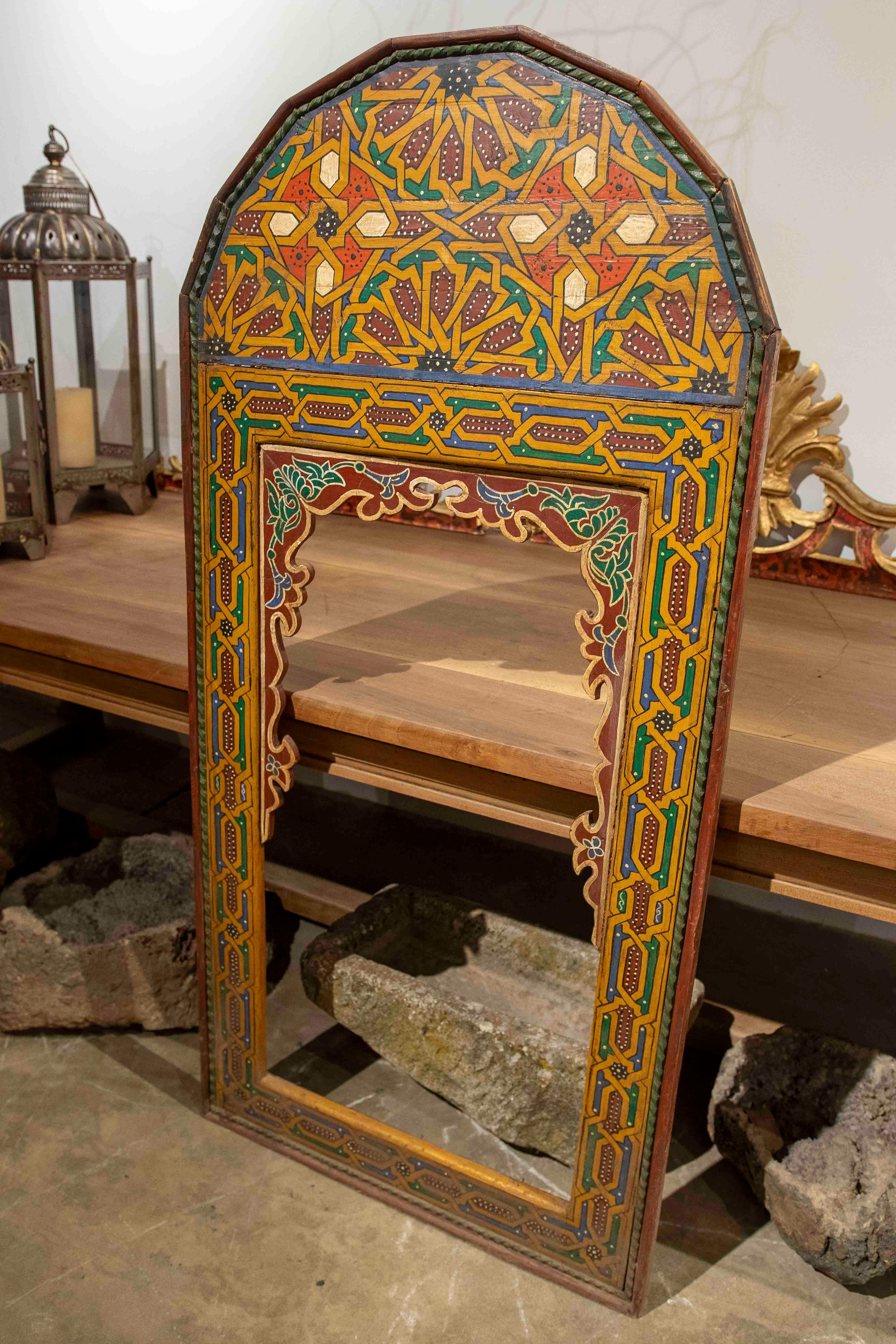 Exquisite hand-painted, polychrome Moroccan mirror with a wooden frame adorned with meticulously crafted geometric and floral motifs. The vibrant colors—reds, greens, yellows, and blues—evoke the rich artisanal traditions of North Africa, where
